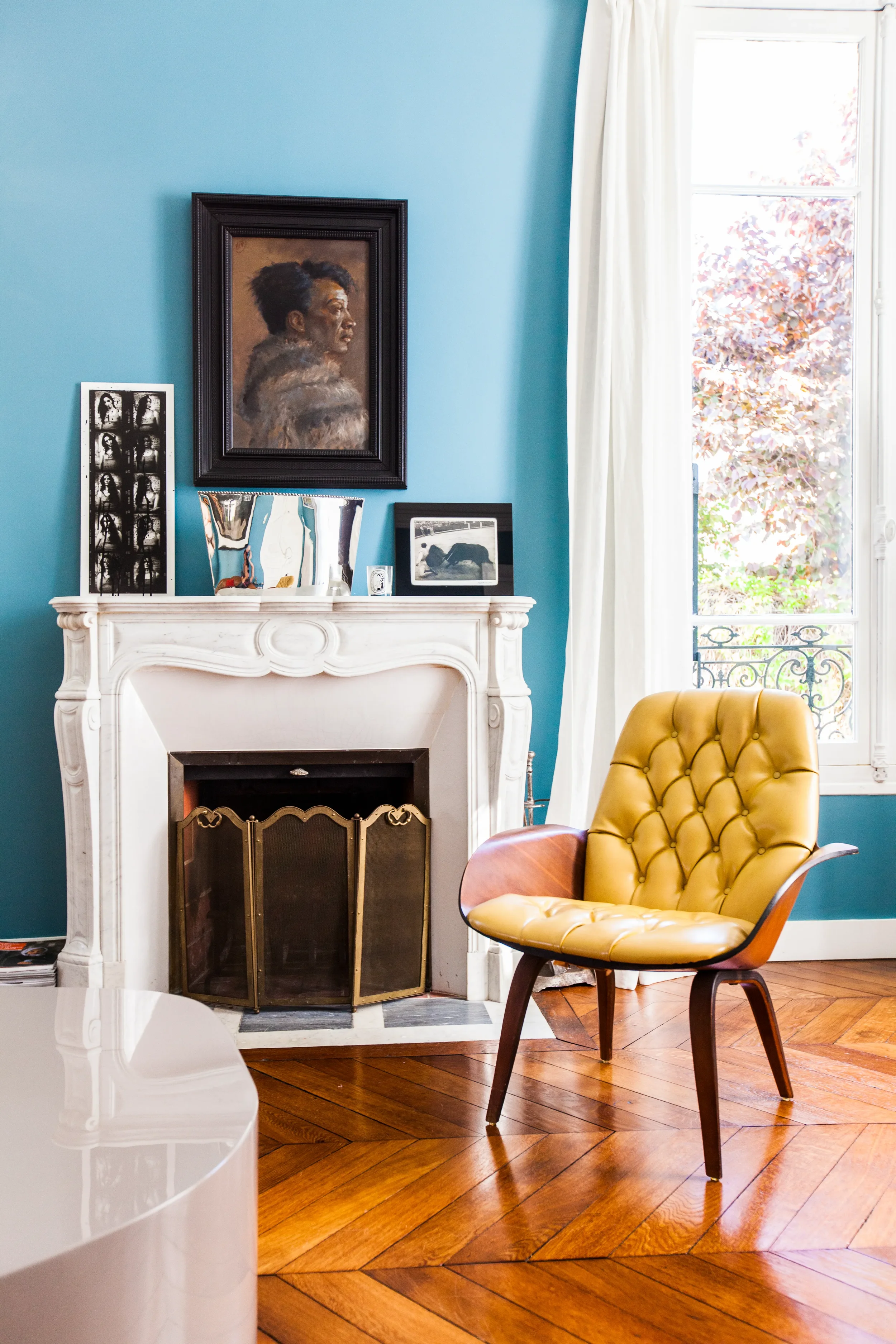 Salon élégant au mur bleu avec cheminée ancienne en marbre blanc, fauteuil capitonné en cuir jaune mustard et parquet en point de Hongrie