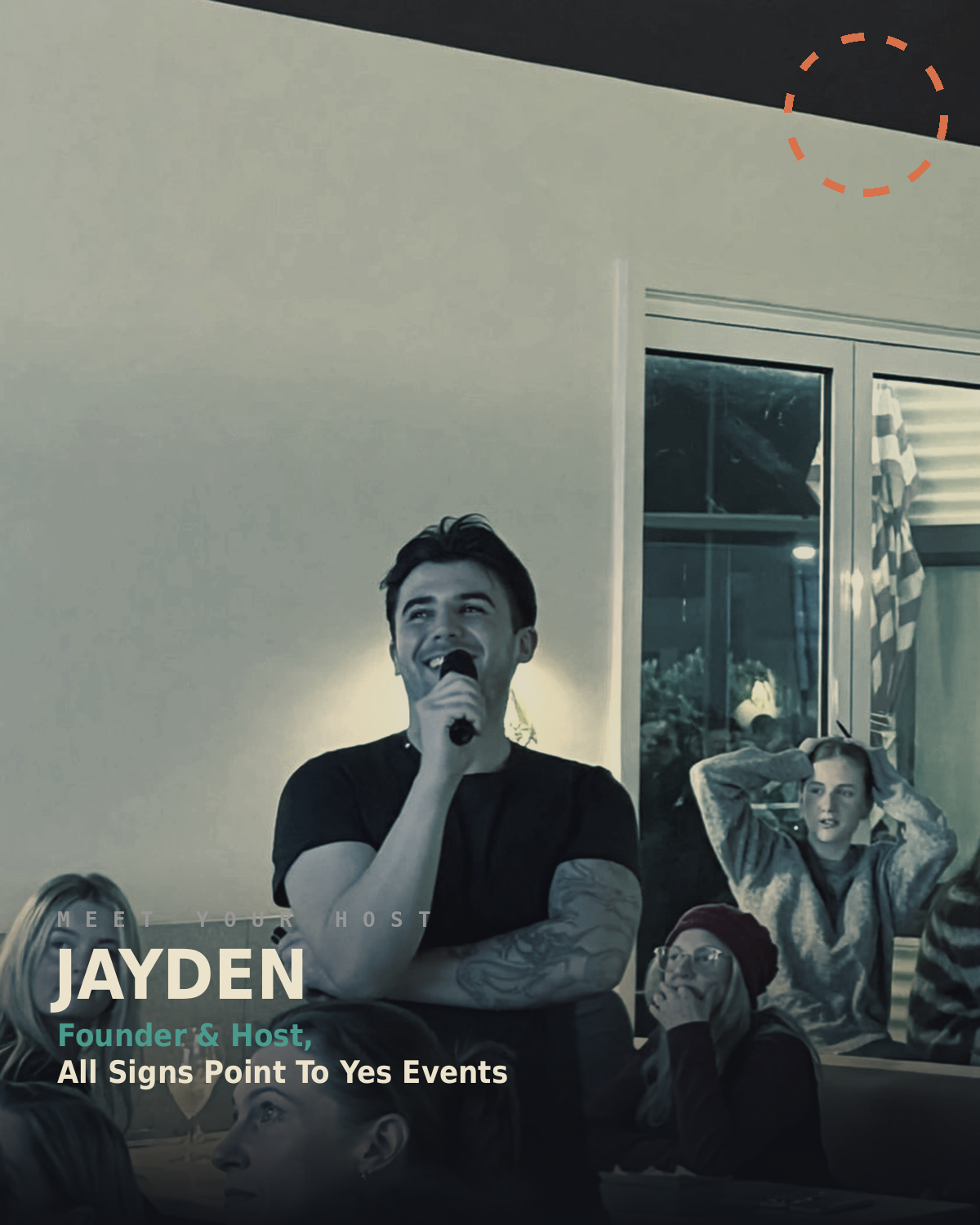 A young man is holding a microphone and smiling during a presentation or speech in a room with a group of people. There is a woman in the background with her hands on her head, and several other people are seated, looking toward the speaker.