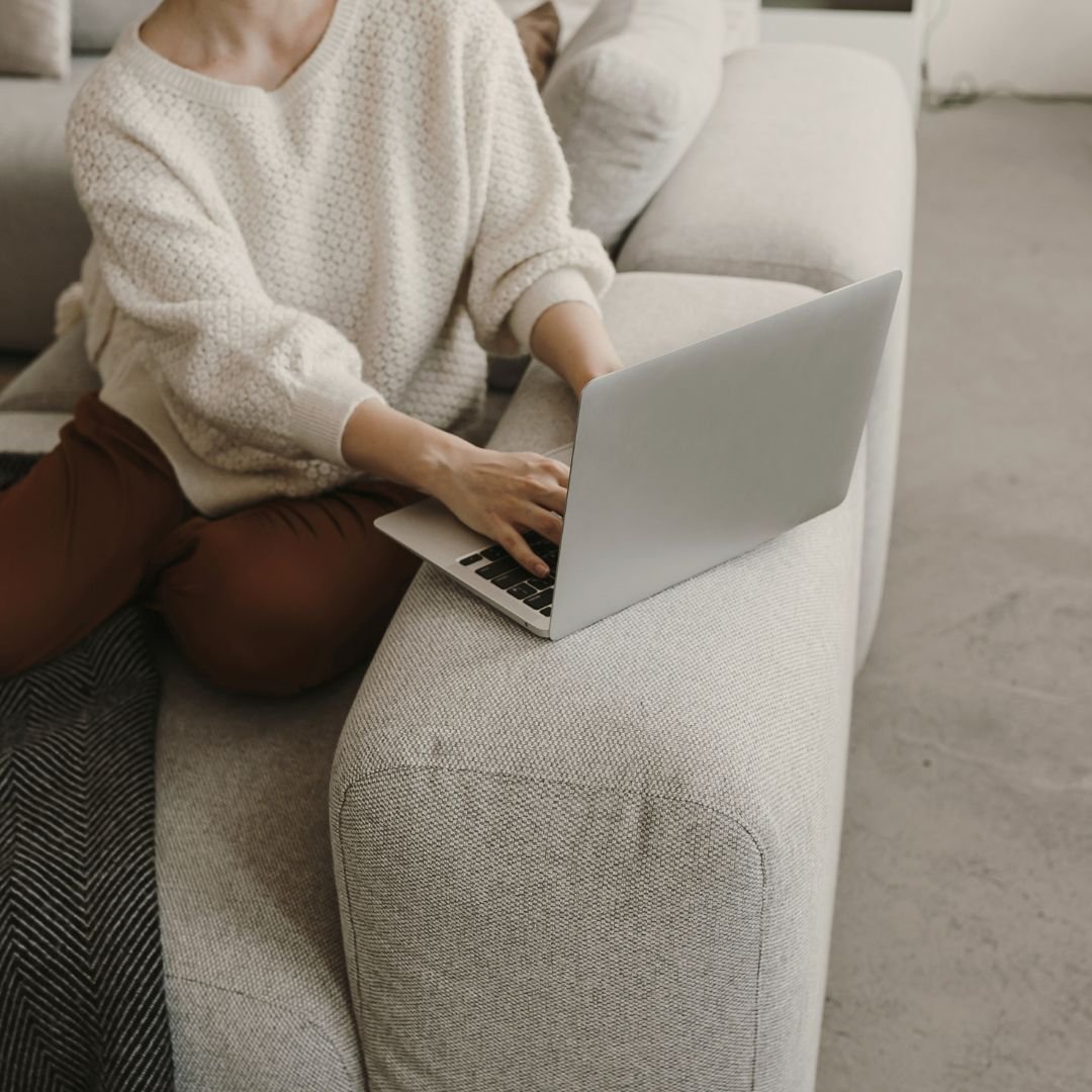 Person sitting on a light-colored sofa using a silver laptop.