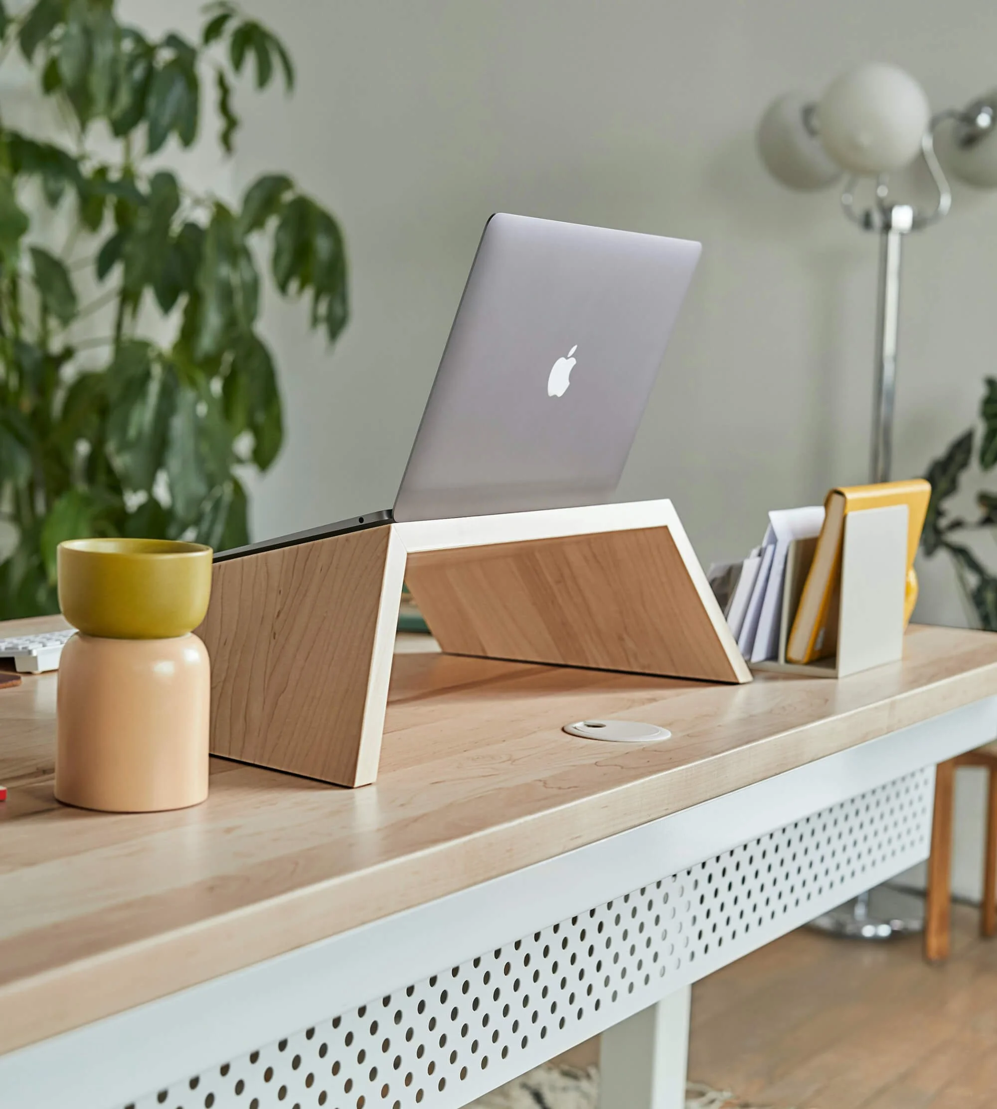 A wooden desk with a laptop stand, holding a closed MacBook, a yellow and beige ceramic cup, a yellow notebook, and some papers, with a large green plant and a floor lamp in the background.