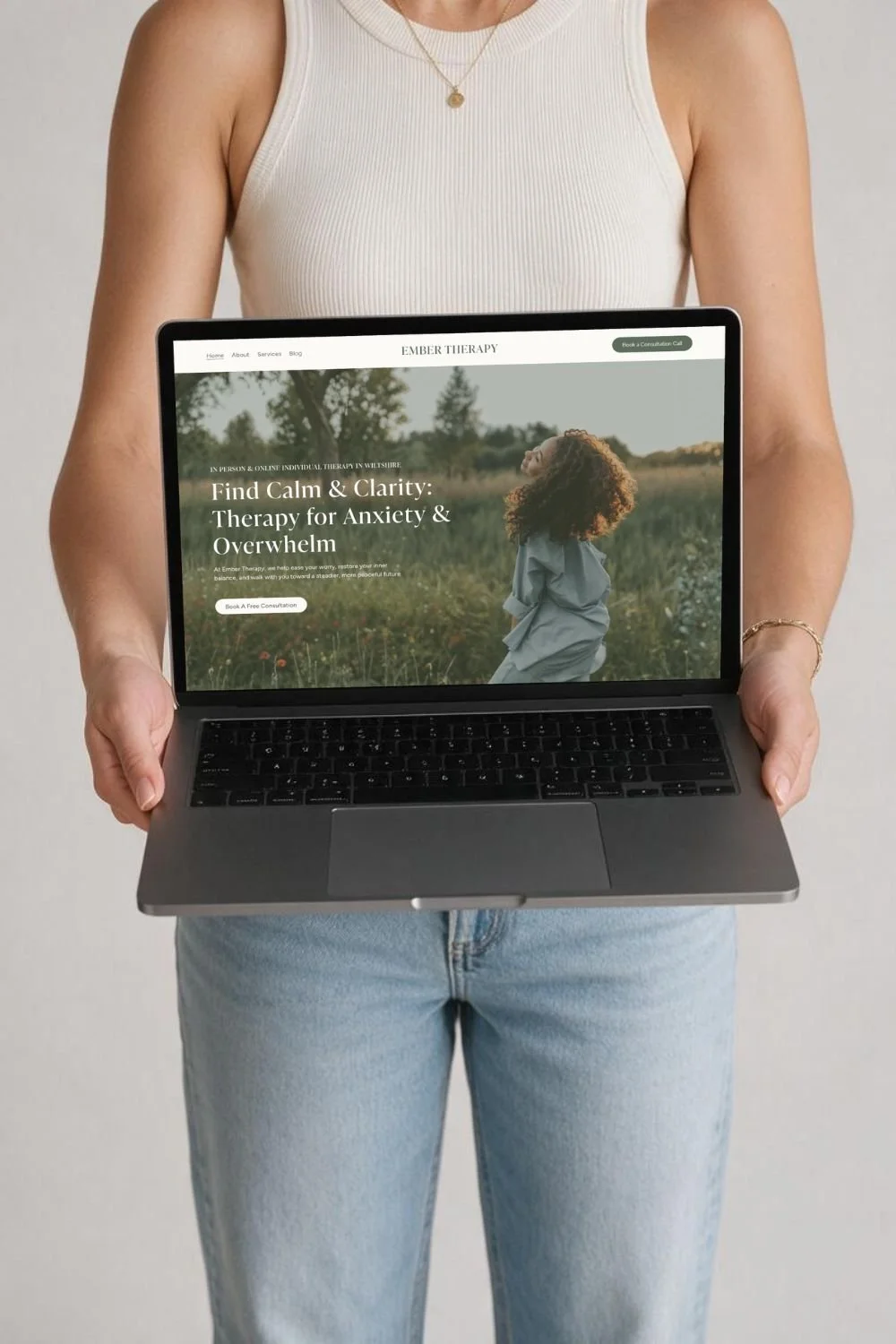 Person wearing a white tank top and light blue jeans holding a laptop displaying a therapy website for anxiety and overwhelm.