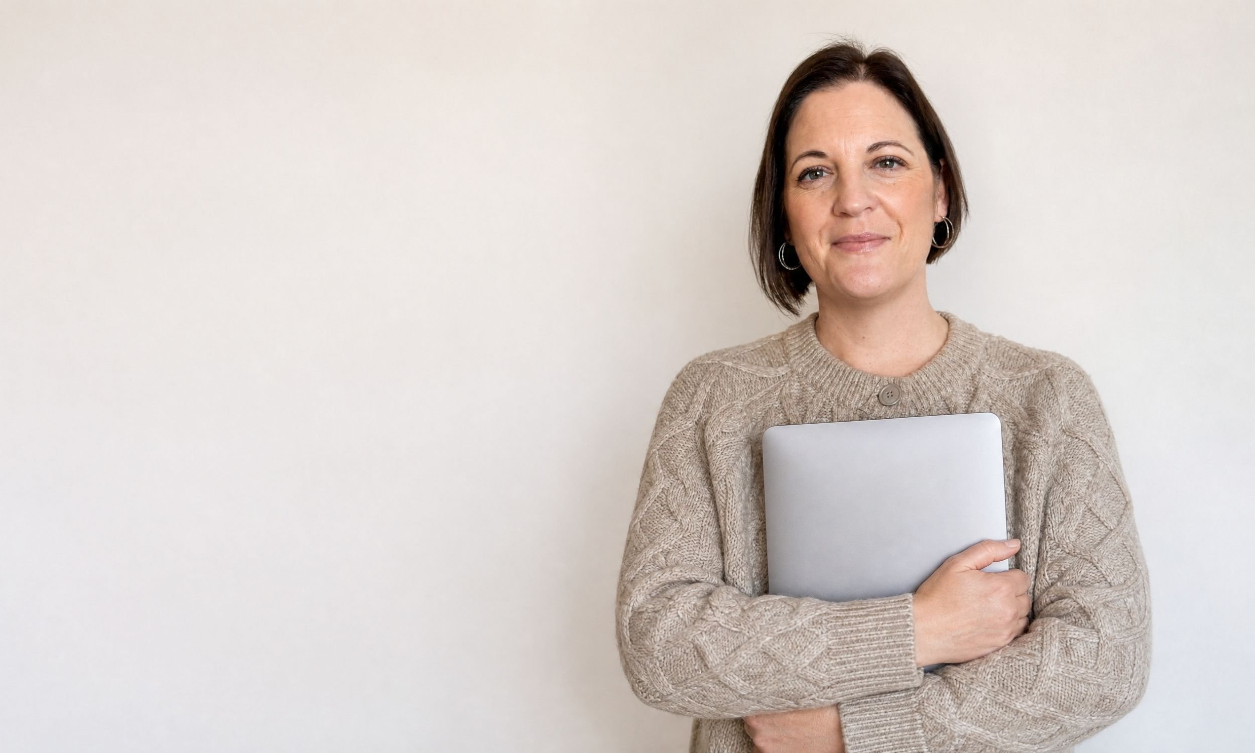 Owner of The Visible Therapist Vicky with short dark hair and hoop earrings, wearing a beige sweater, holding a silver laptop, standing against a plain white wall.