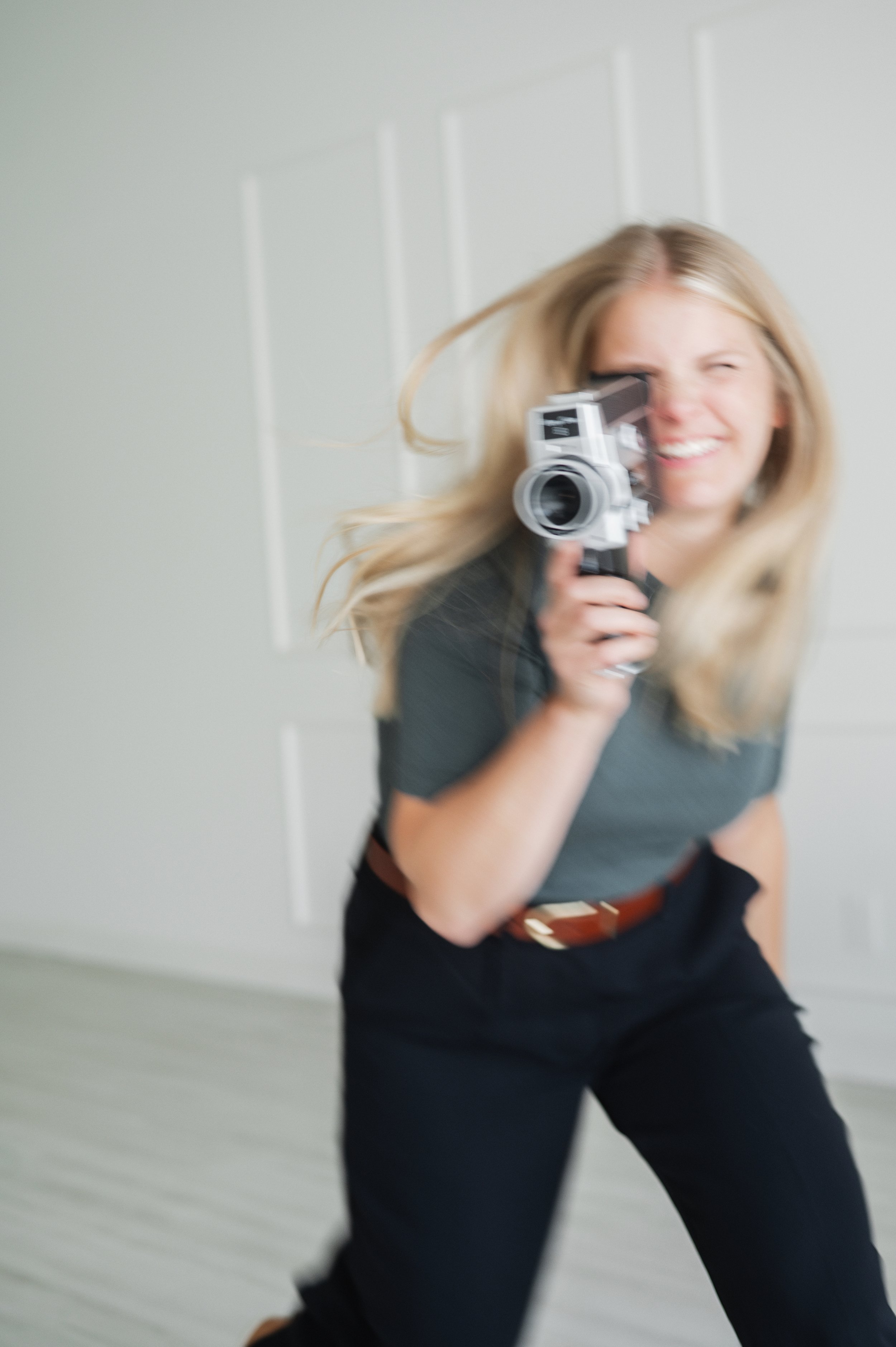 A smiling woman with blonde hair holding a movie camera, pointing it directly at the viewer, in front of a white wall with paneling.