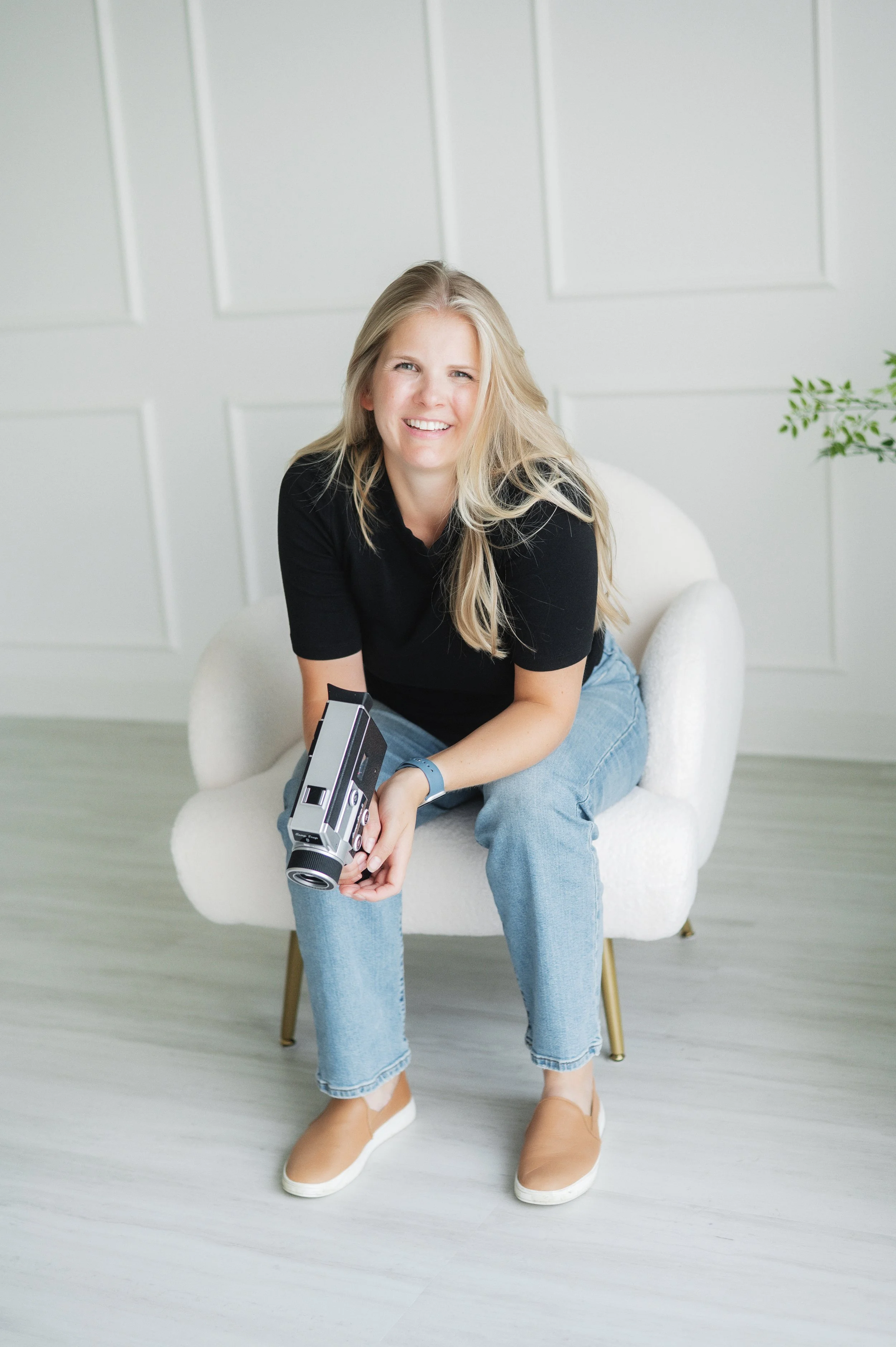 A smiling blonde woman wearing a black shirt and jeans, sitting on a white chair in a bright white room with minimal decor, holding a vintage film camera.