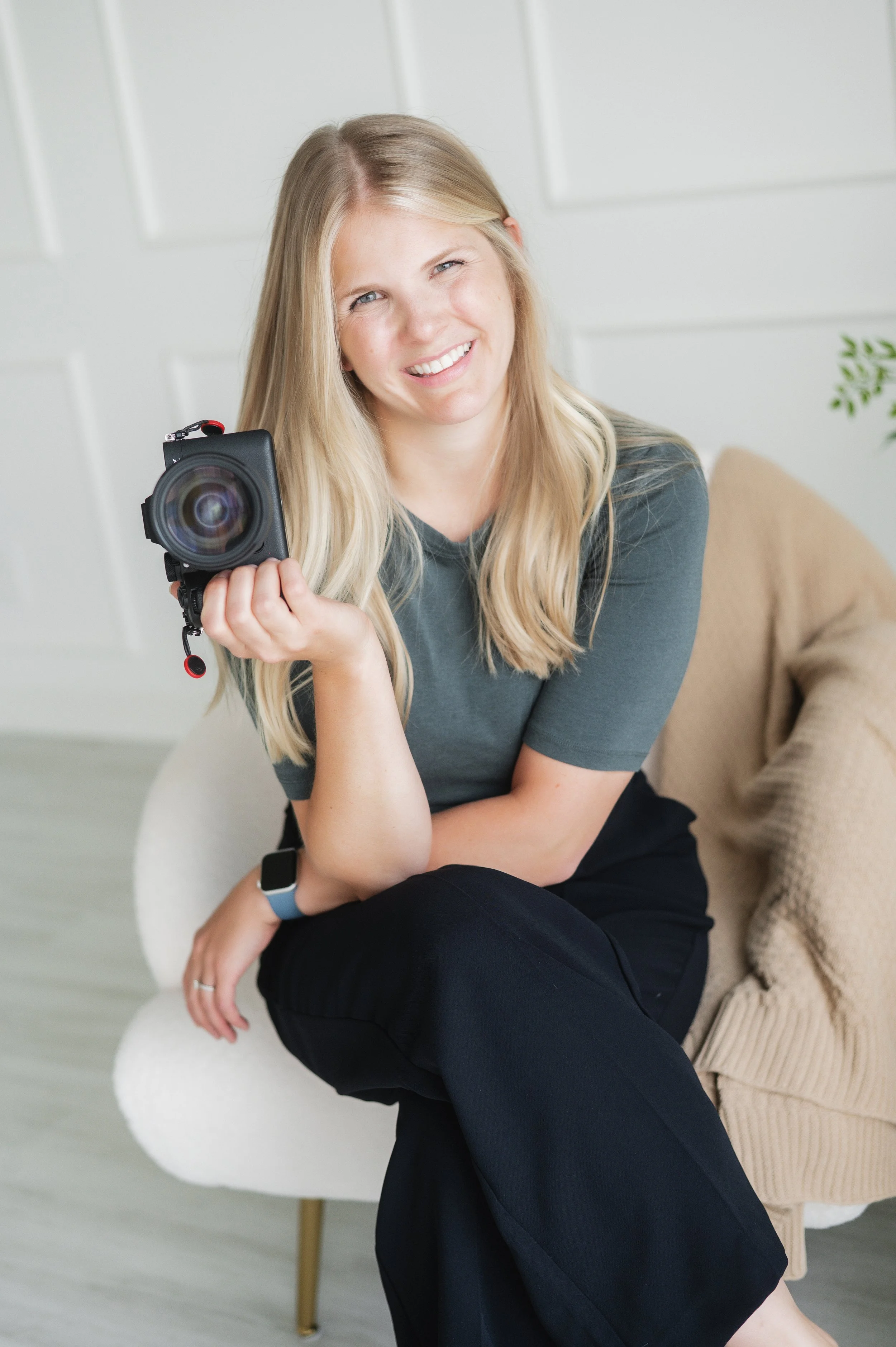 A young woman with blonde hair sitting on a beige armchair, smiling and holding a camera.