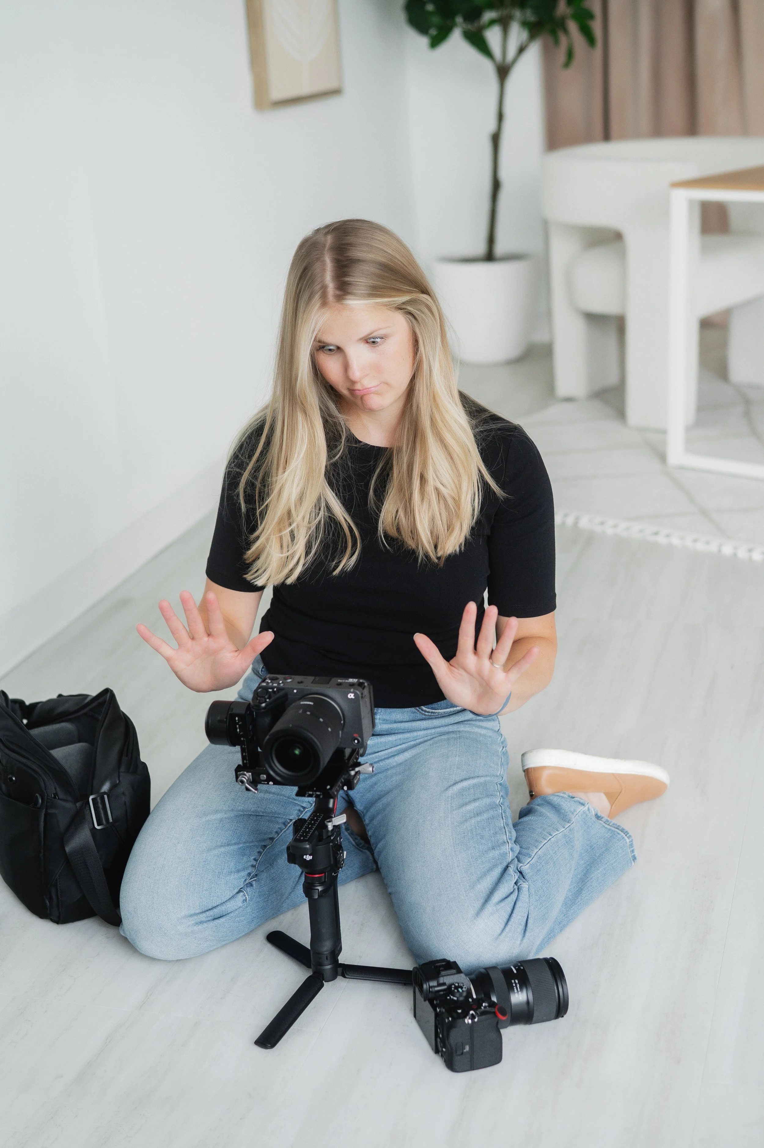 A woman with long blonde hair, wearing a black shirt and light blue jeans, sitting on the floor with a man in similar clothing, adjusting a camera on a tripod. The woman is looking at the camera and holding her hands up. The scene takes place in a bright room with white walls, a potted plant, and some furniture.