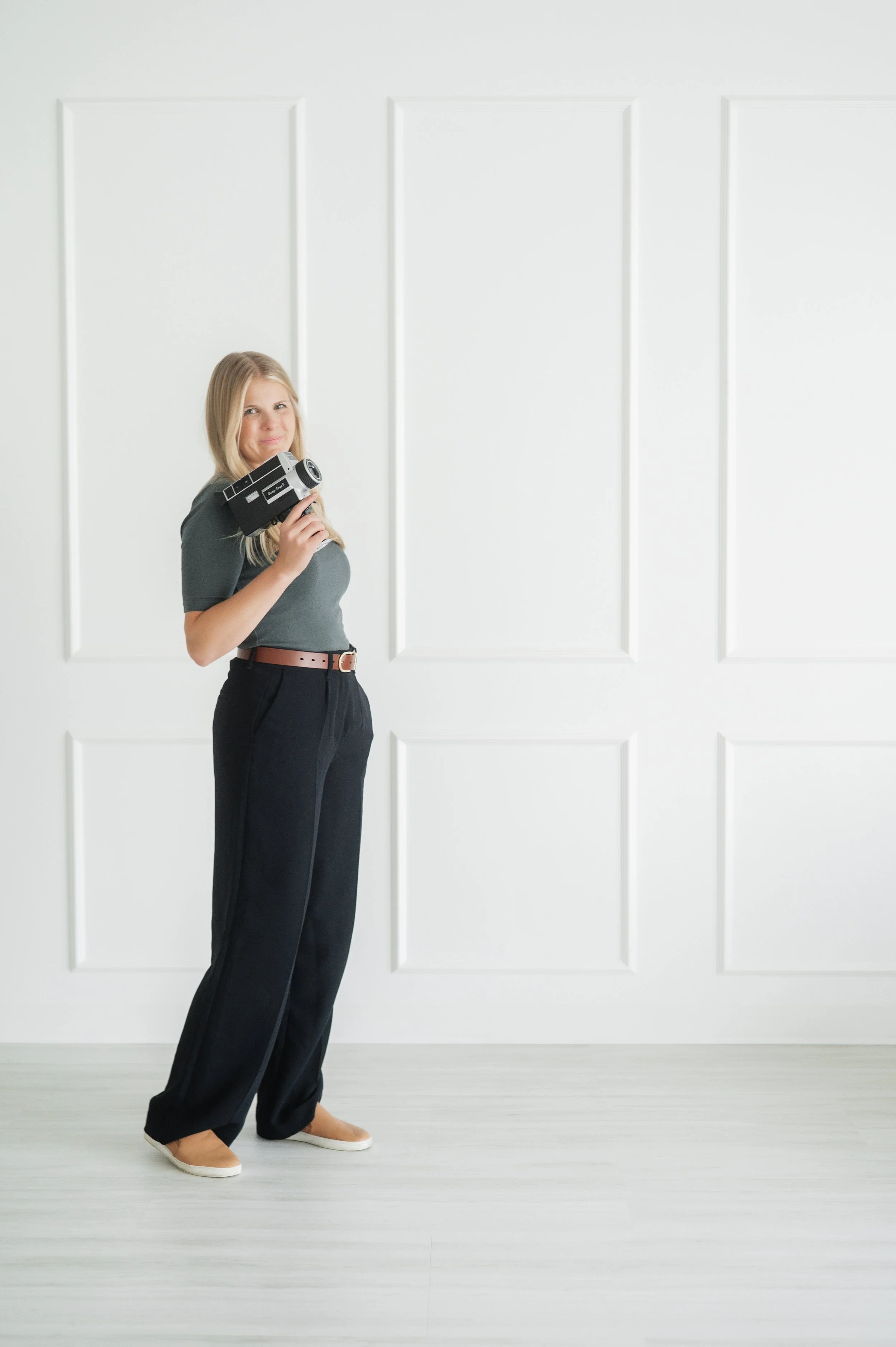 A woman holding a vintage camera standing in front of a white wall with decorative rectangular panels. She is wearing a gray top, black high-waisted pants with a brown belt, and beige slip-on shoes.