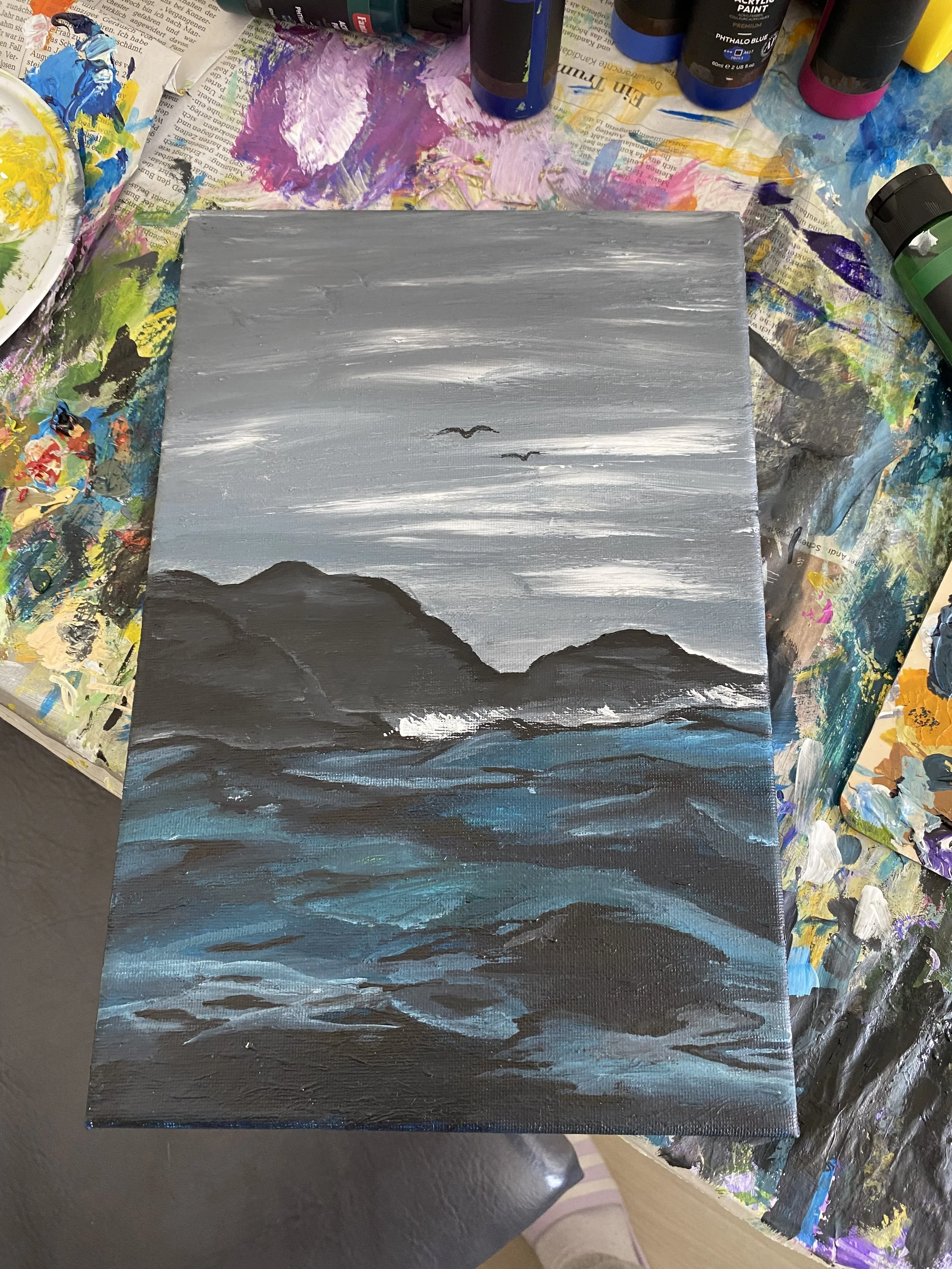 A landscape painting in shades of gray, black, and blue, depicting mountains and a cloudy sky, created on a canvas.