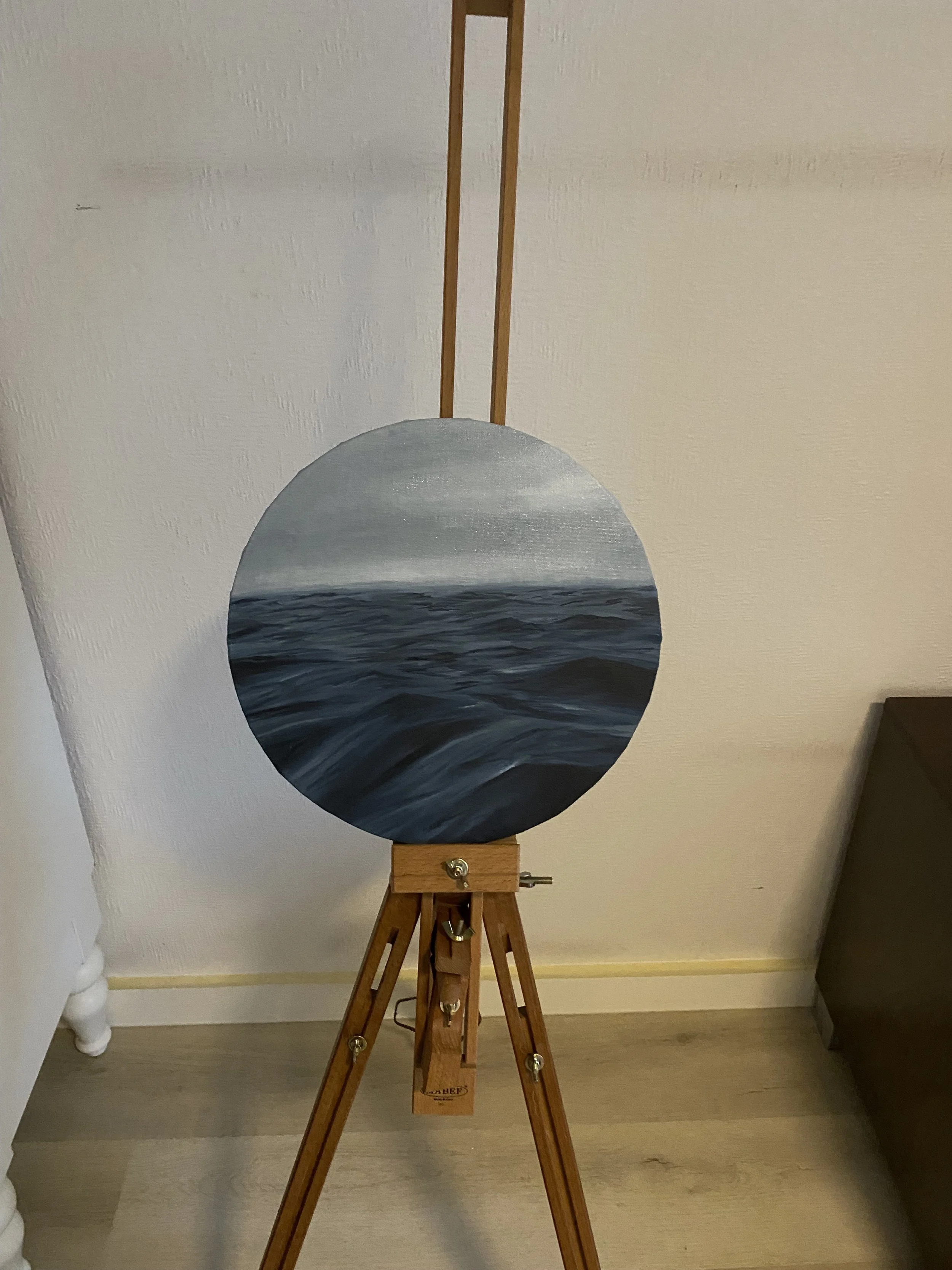 Ein Holzeasel mit einer runden, gemalten Scheibe, die ein Meer mit Wellen zeigt, ist in einem Raum aufgestellt.