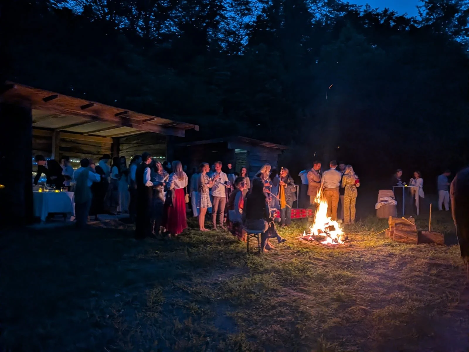 Guests gather around a campfire at night during an outdoor event, with some standing and others sitting, near a wooden shelter, under a dark sky with tall trees in the background.