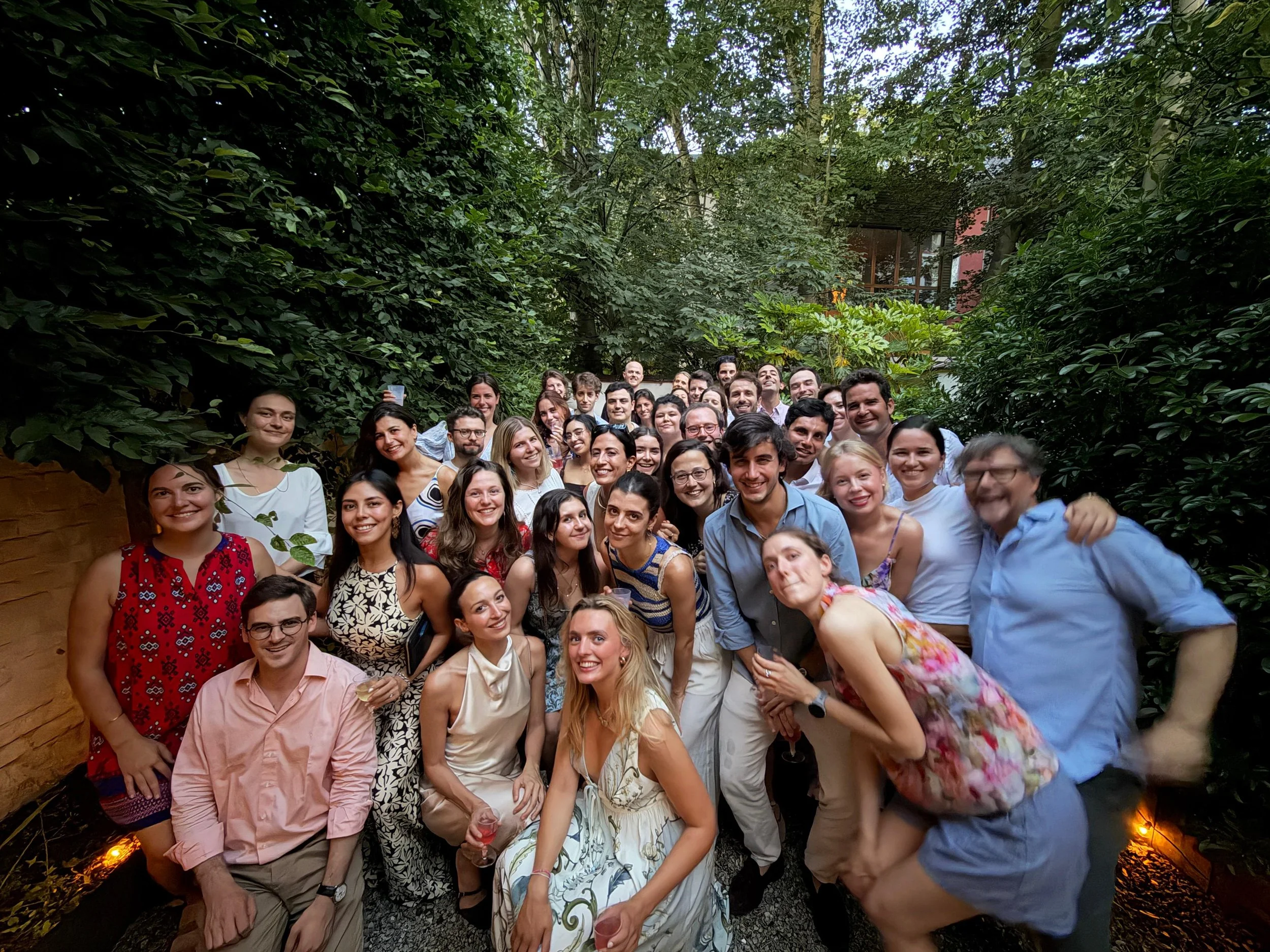 Group of people gathered outdoors for a celebration, smiling, surrounded by lush green foliage, some holding drinks.