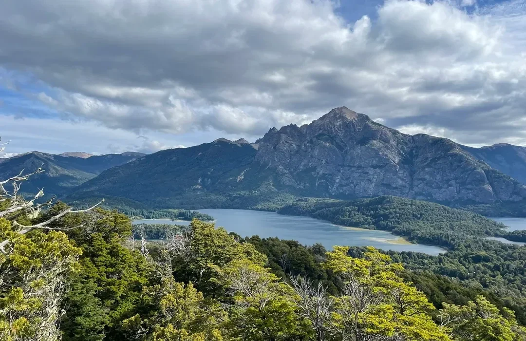 A panoramic view of a mountain range with a lake in the foreground, forests surrounding the lake, and partly cloudy skies.