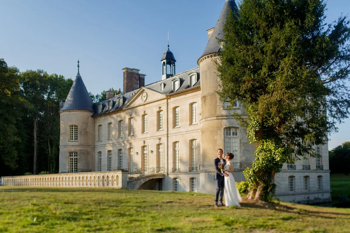 A newlywed couple standing near a large castle with a tower and turrets, surrounded by green trees, on a sunny day.