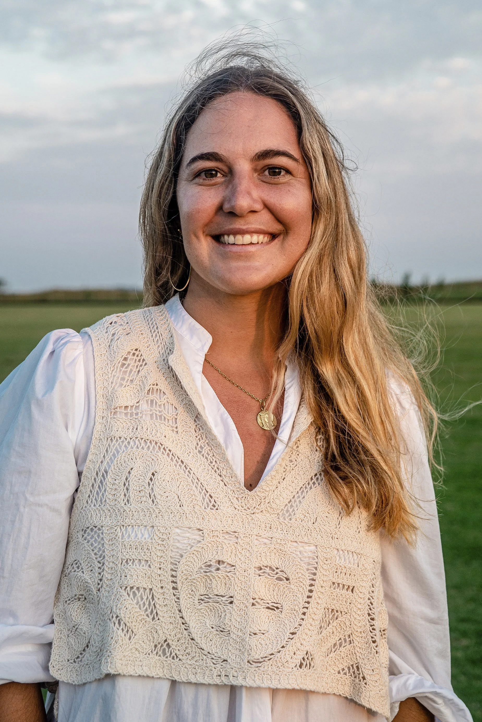 Close-up of a smiling woman with long wavy hair outdoors, wearing a white blouse and a beige crocheted vest.