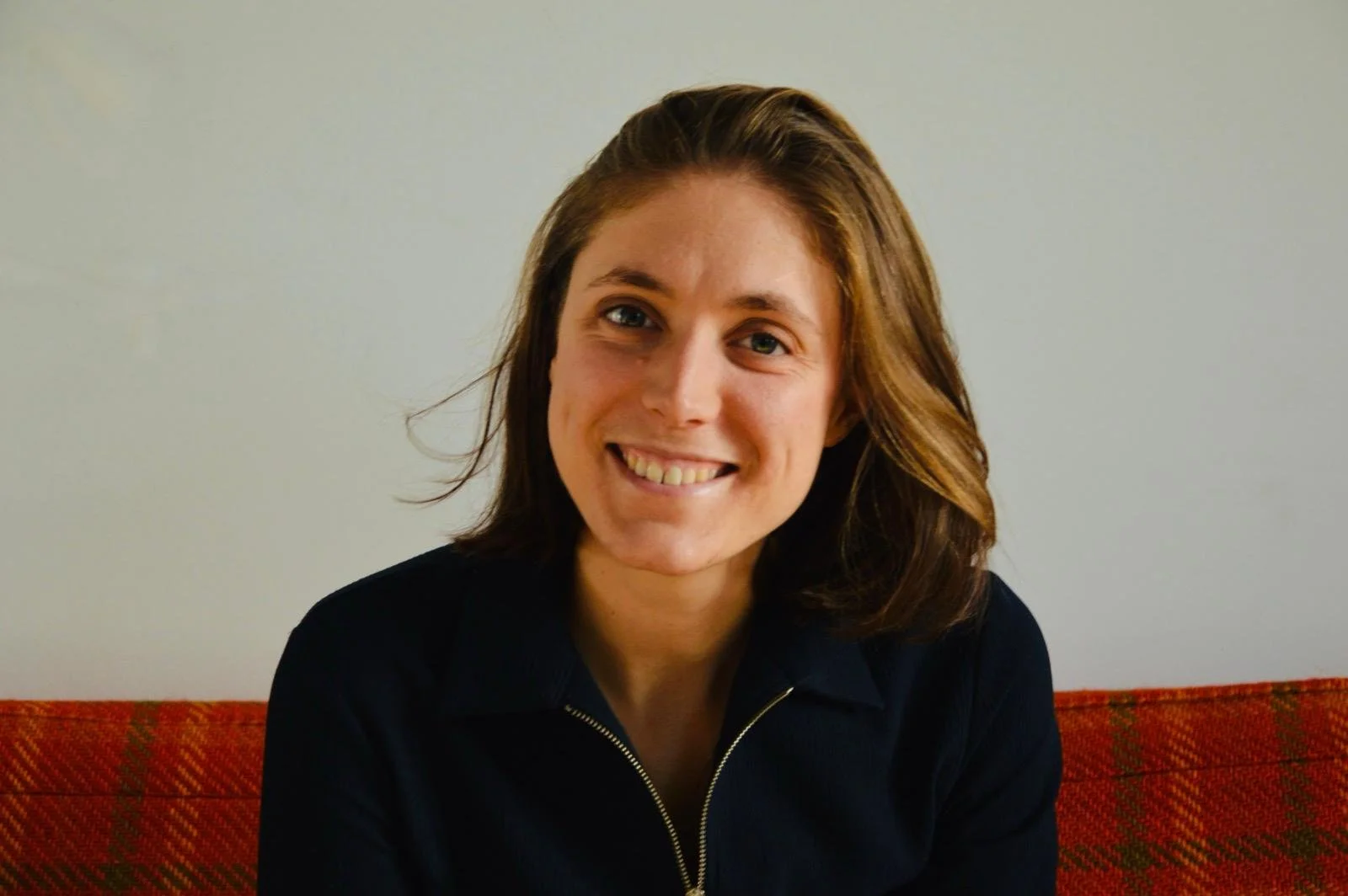 A woman with medium-length brown hair smiling, wearing a dark top, seated on a red and orange patterned couch against a plain light-colored wall.