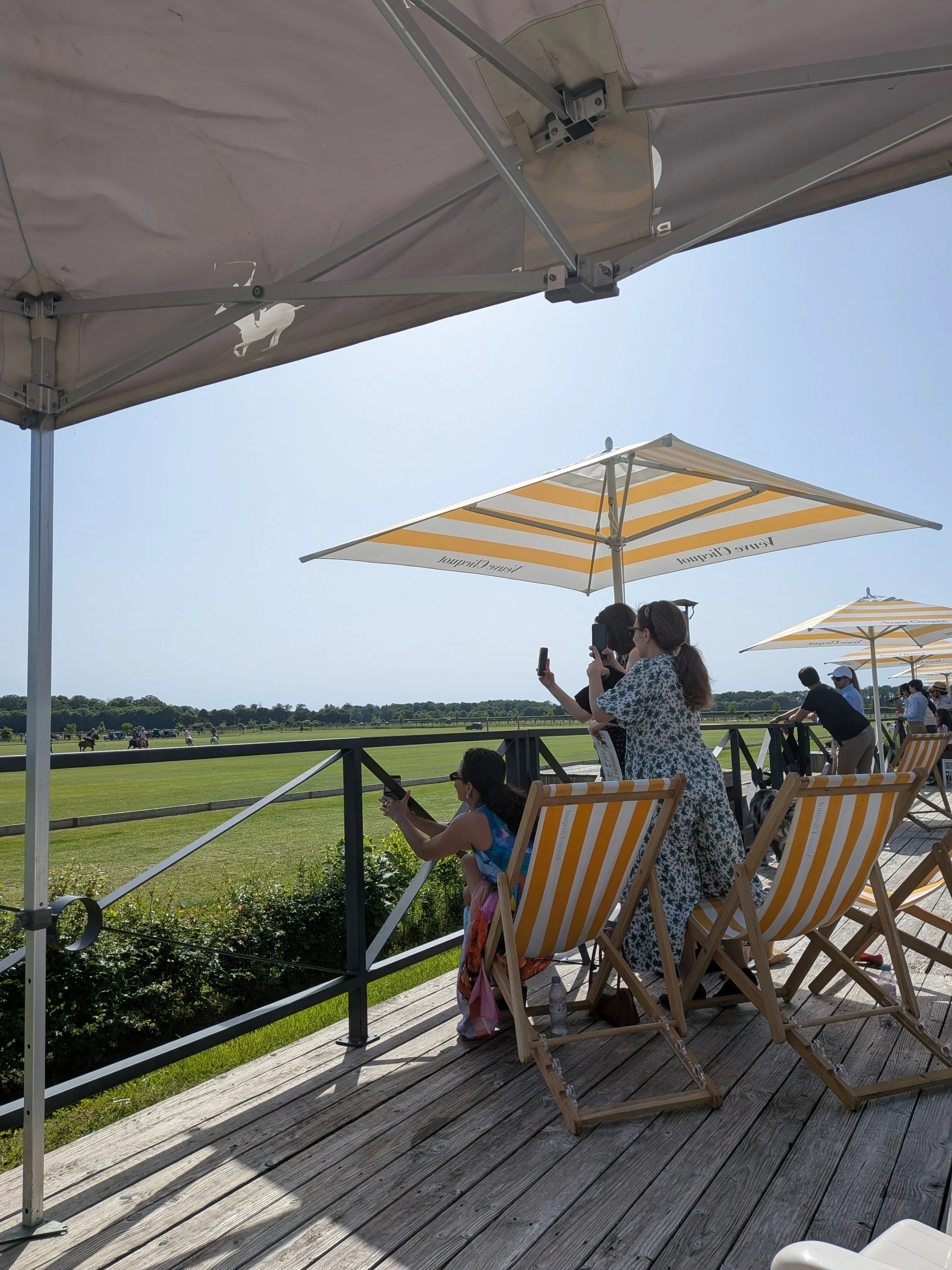 People sitting on wooden deck chairs under yellow-and-white striped umbrellas at a horse racecourse. Some are taking photos, others are looking at the race in the background on a sunny day with clear blue sky.