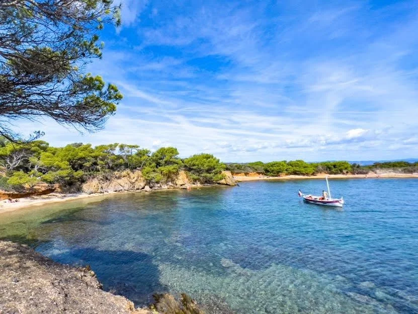 A peaceful cove with clear blue water, a small sailboat, and a rocky shoreline lined with green trees under a partly cloudy sky.