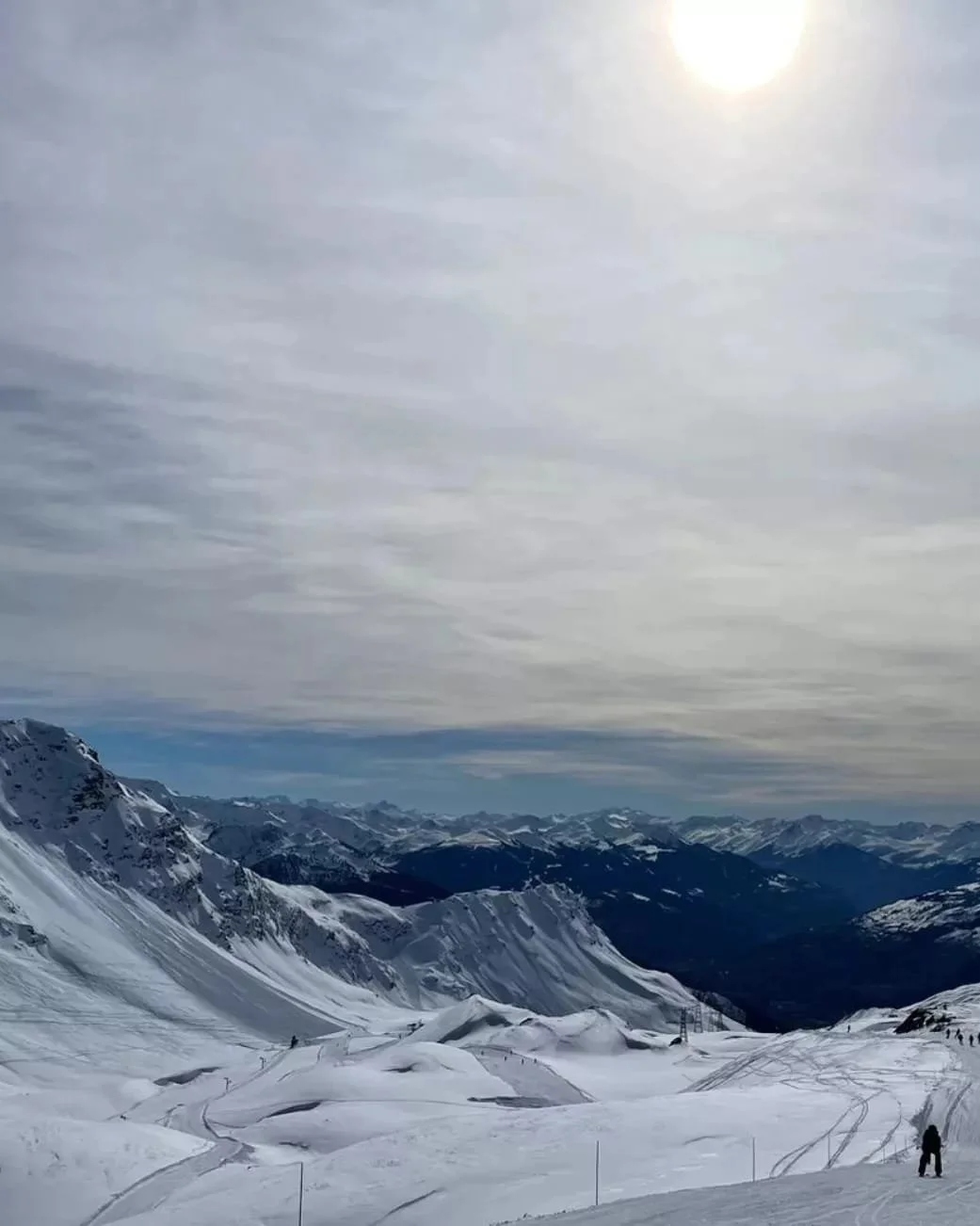 Snow-covered mountain landscape with ski tracks and a skier, cloudy sky, and the sun faintly visible.