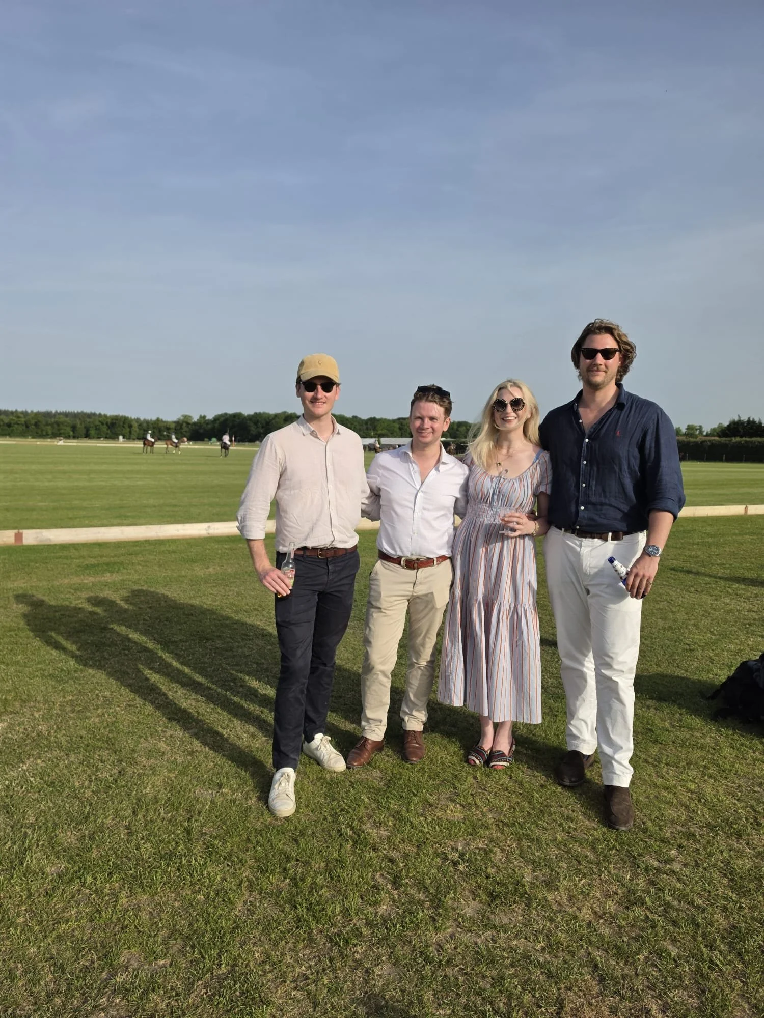 Four people standing on a tennis field on a sunny day, with horses and a fenced track in the background.