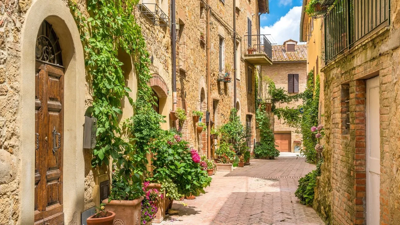 A narrow cobblestone street in a European village with old stone buildings, plants, and flowers along the sides, under a partly cloudy sky.
