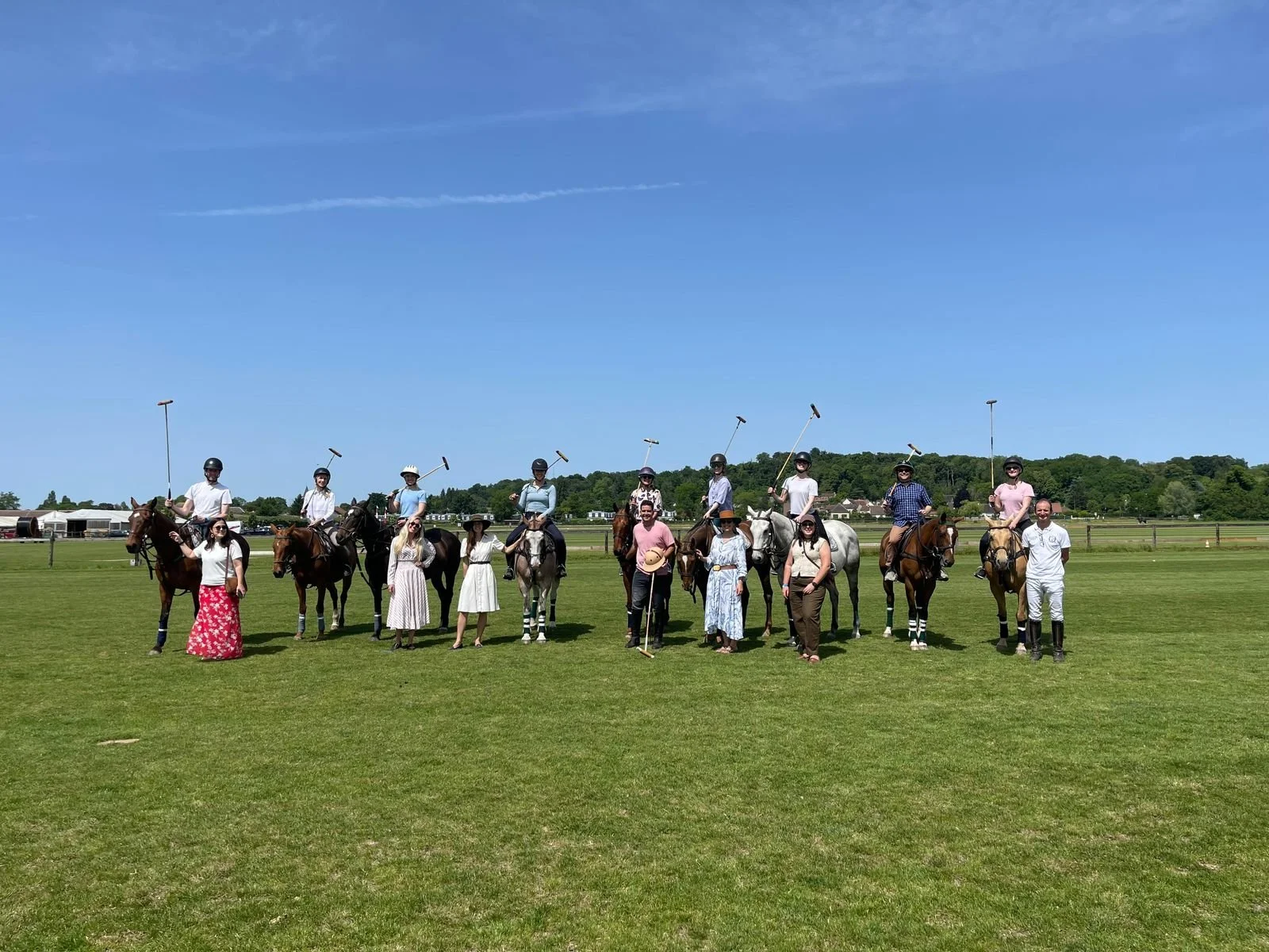A group of people on horseback and standing on green grass at a polo field, with a clear blue sky overhead.