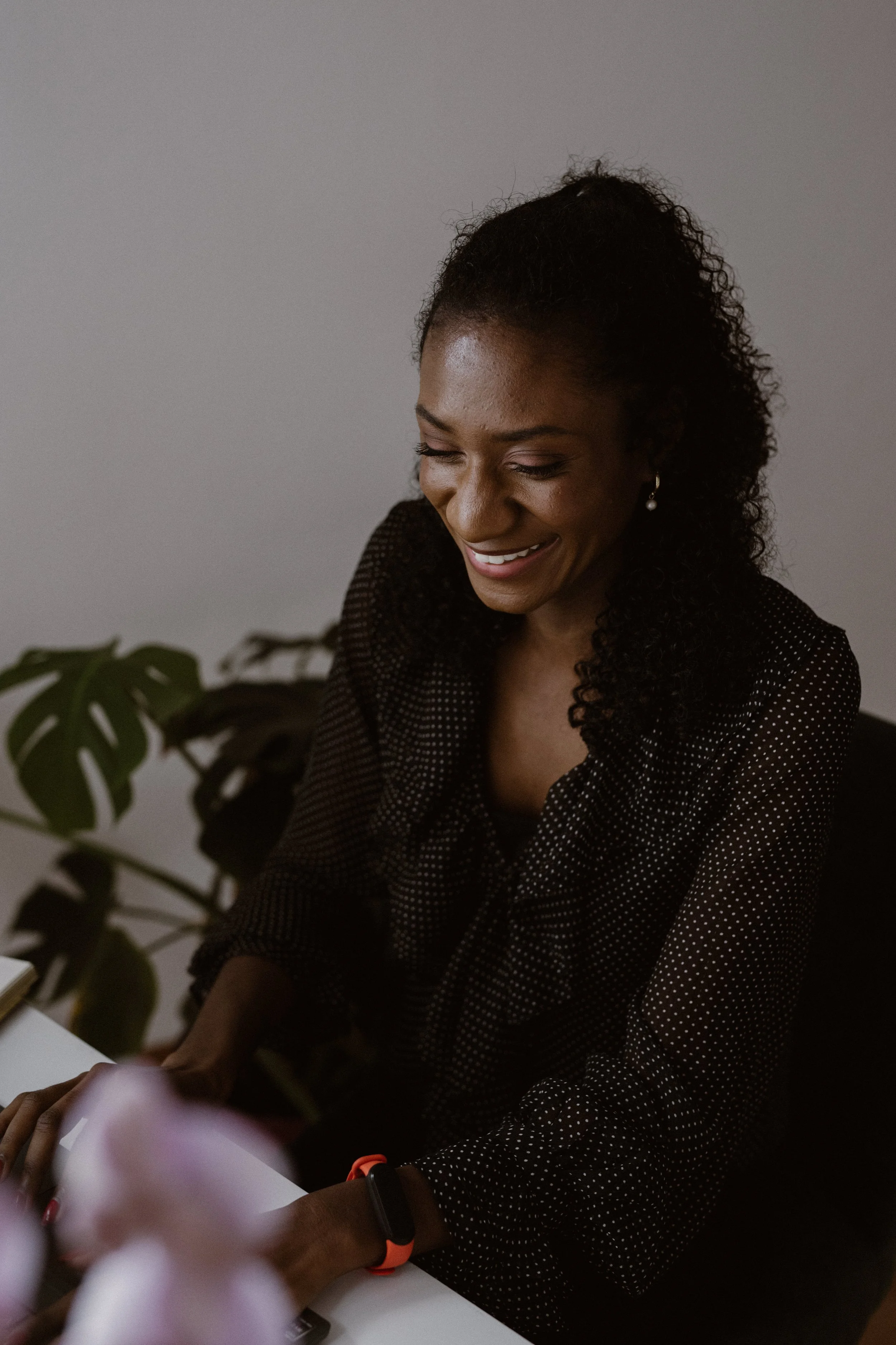 A woman with curly hair smiling and looking down, wearing a black polka dot top and pearl earrings, sitting at a table with a plant nearby.
