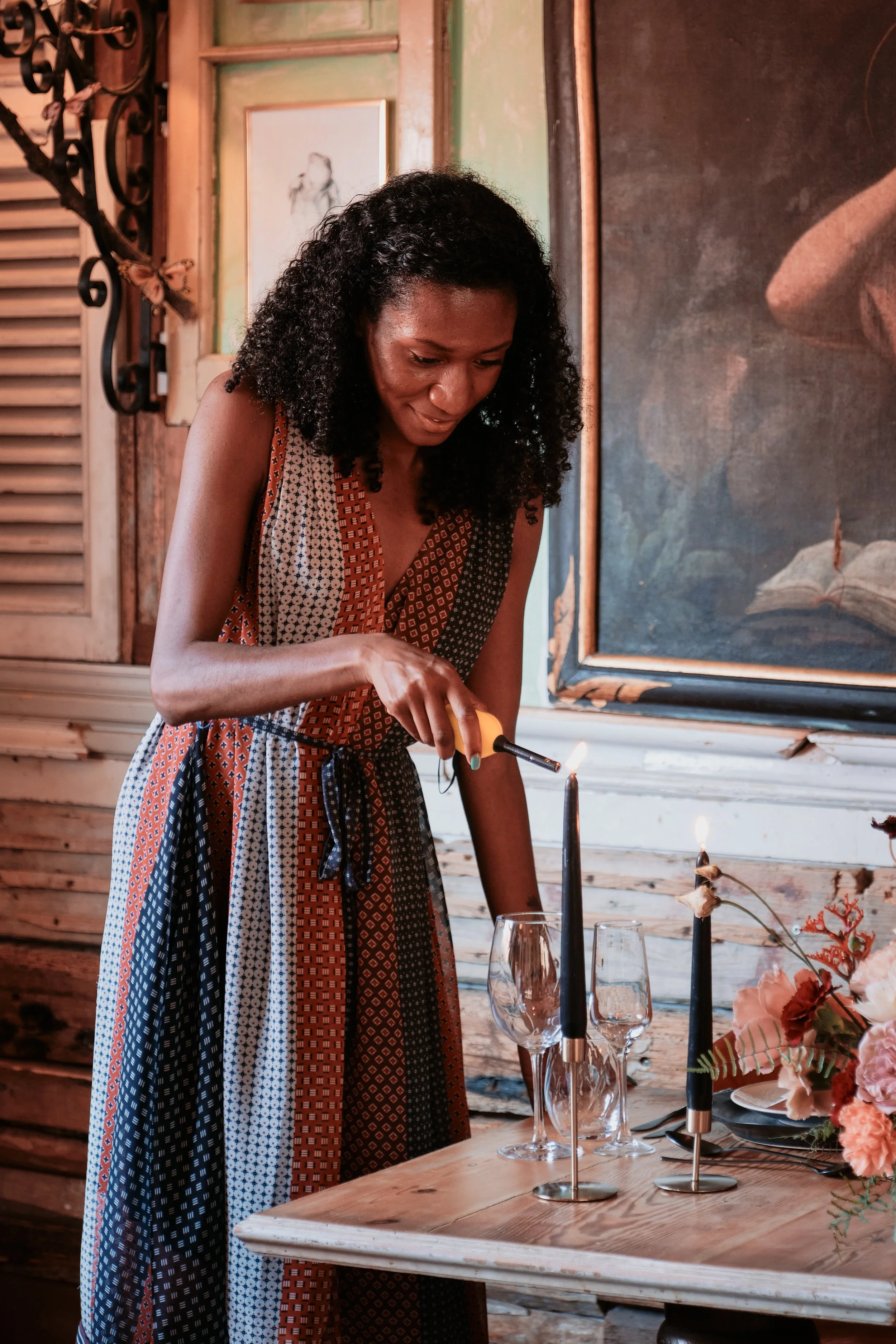 A woman with curly black hair in a patterned dress is lighting a candle at a table set with glasses, flowers, and a black candle holder, in a rustic wooden room.