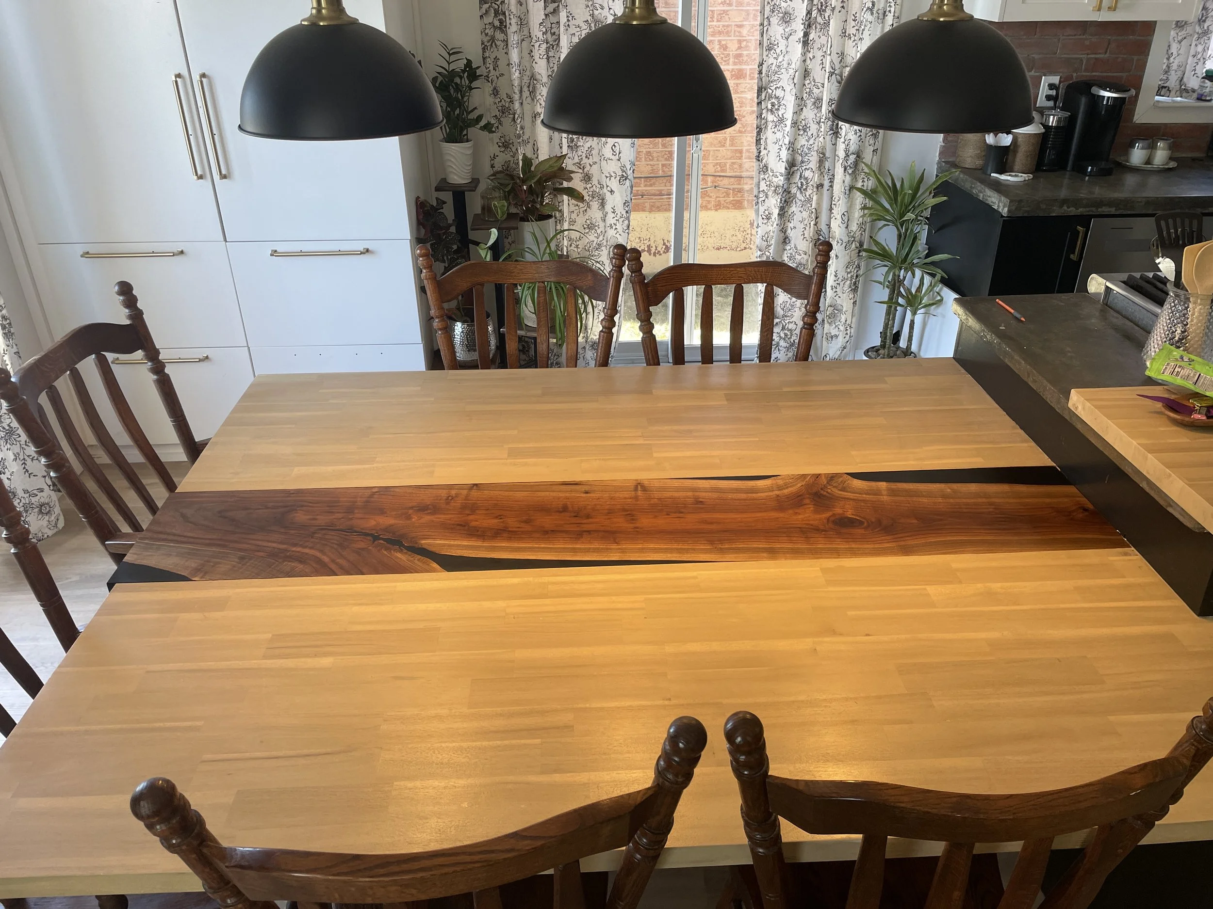 Wood dining table with dark wood chairs in kitchen, black pendant lights hanging above, plants near window with floral curtains, brick wall and kitchen appliances in background.