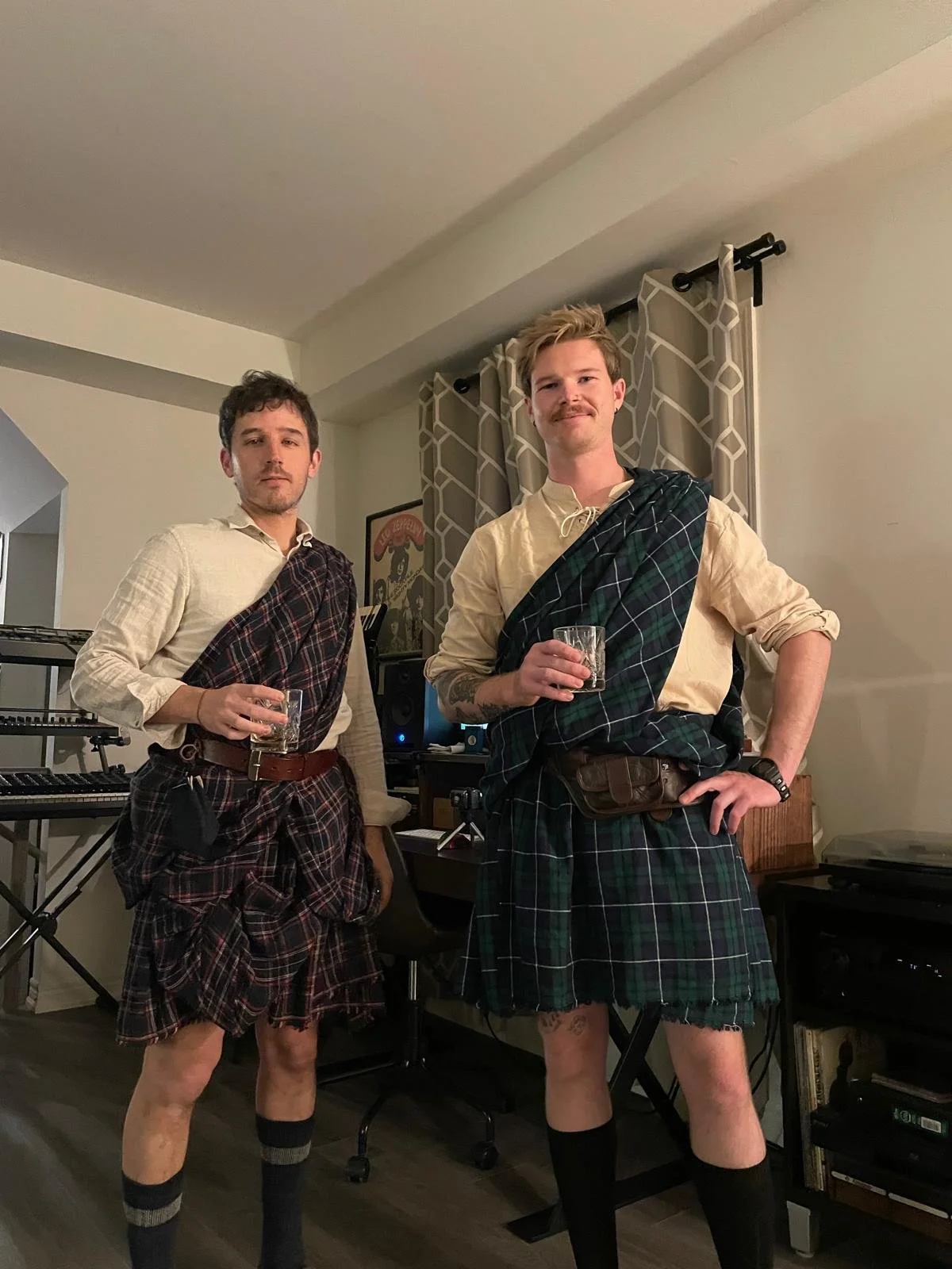 Two men standing indoors holding glasses of whiskey, wearing traditional Scottish kilts and shirts, with a music keyboard and studio equipment in the background.