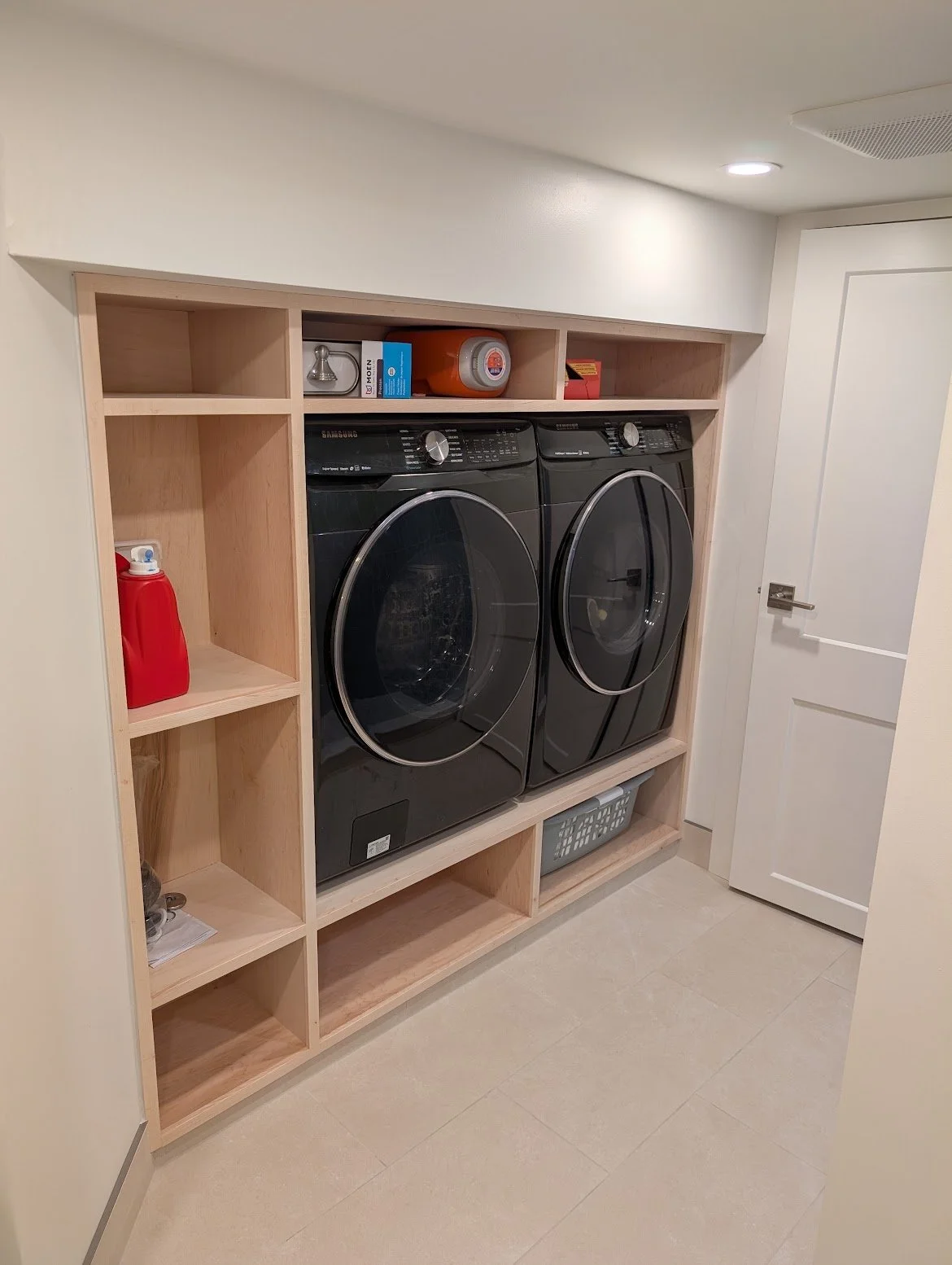 A laundry room featuring a built-in wooden shelving unit with a red detergent bottle, a grey laundry basket, and various small items. Inside the shelving unit are a front-loading washing machine and dryer in black. The room has beige tile flooring, white walls, and a white door.