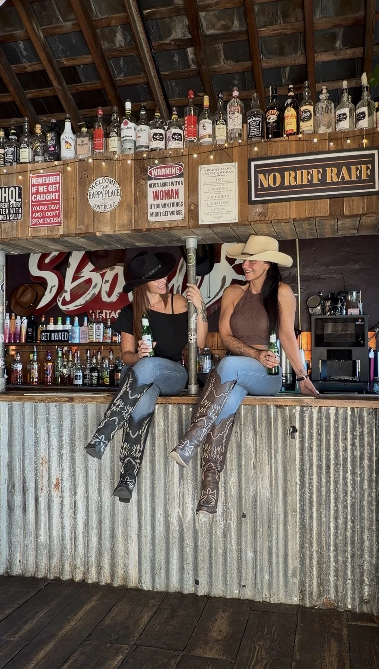 Two women sitting on a bar counter, wearing cowboy boots and wide-brimmed hats, holding bottles of beer, with a rustic bar setup in the background that includes liquor bottles and decorative signs.