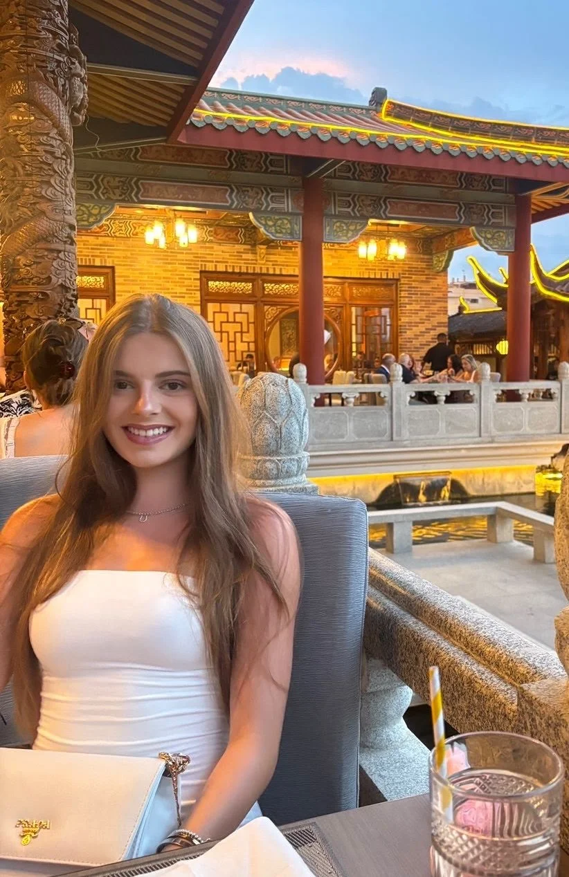 A young woman with long brown hair smiling and sitting at an outdoor restaurant with traditional Asian architectural elements, including ornate wooden carvings, red columns, and a pagoda-style roof, during evening time.