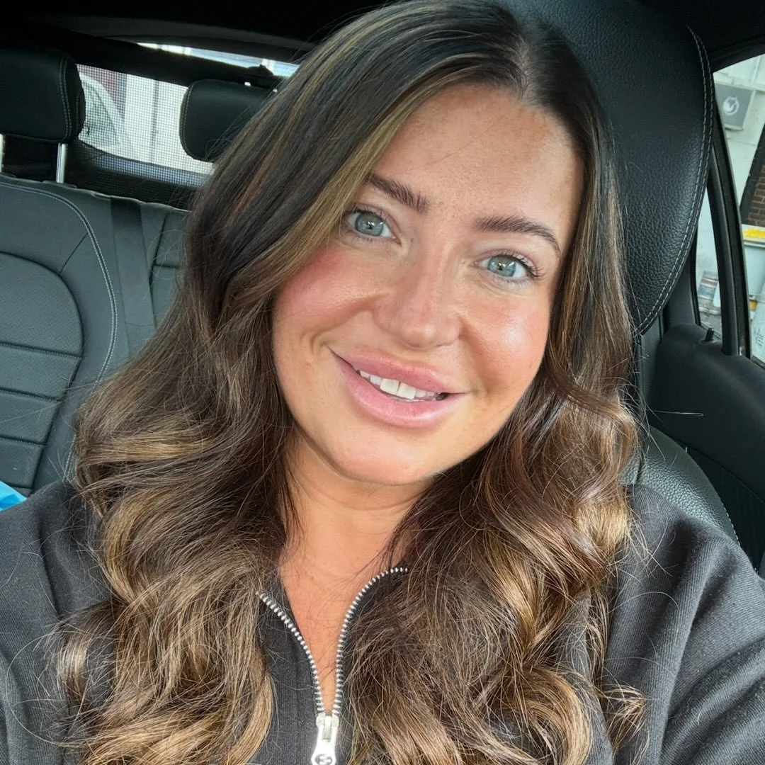 Close-up of a woman with long wavy brown hair and blue eyes, smiling inside a car.