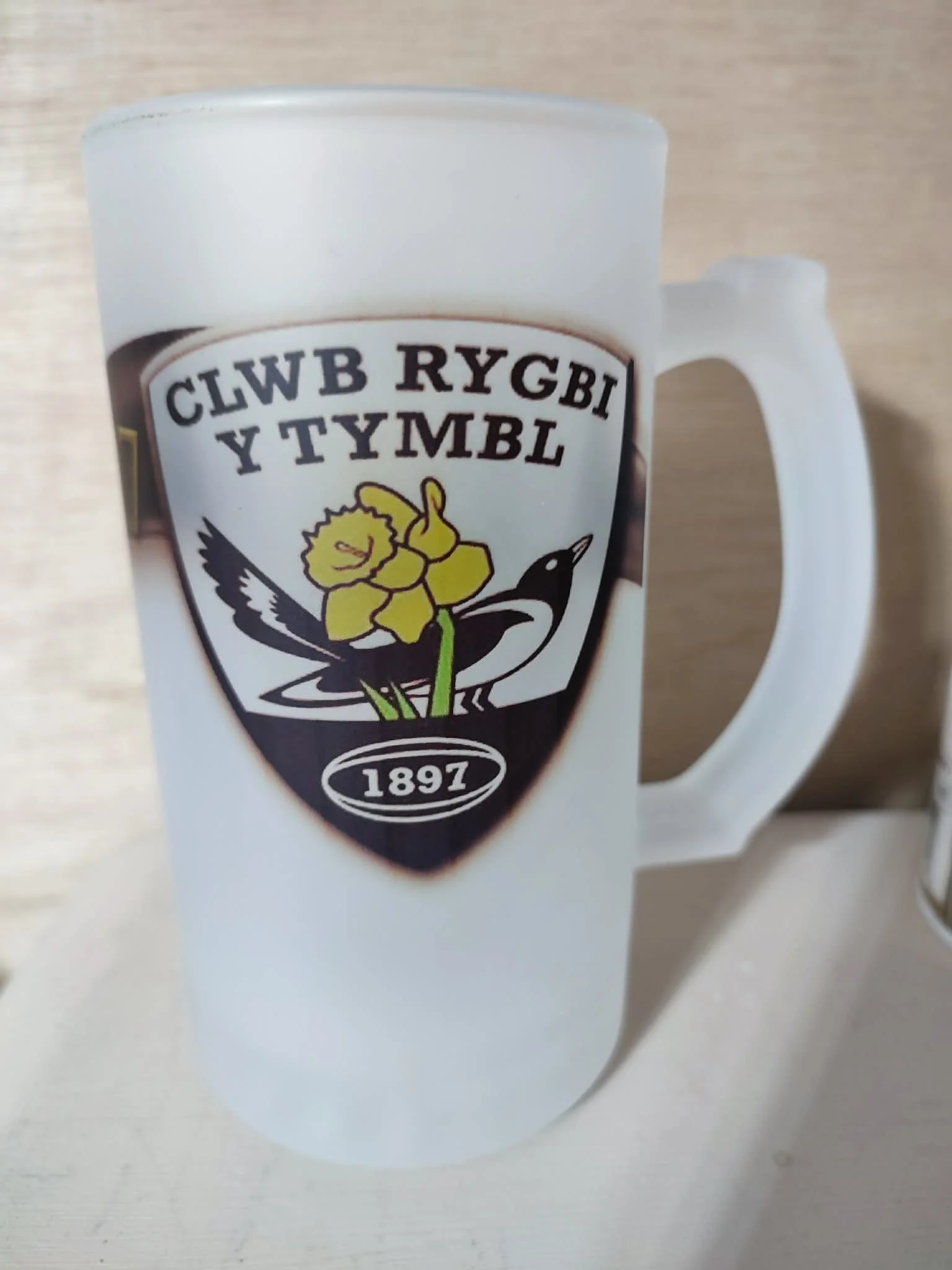 White ceramic mug with a crest logo featuring a yellow flower, a black and white bird, and text saying '1897'.