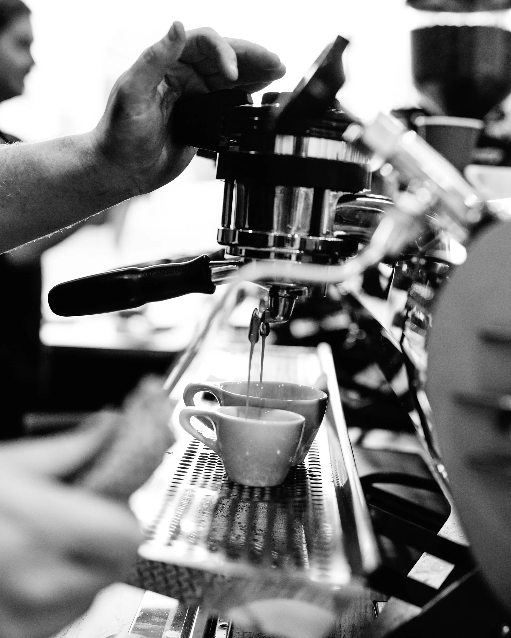 Black and white photo of a person pouring hot espresso into a small cup from a professional espresso machine.
