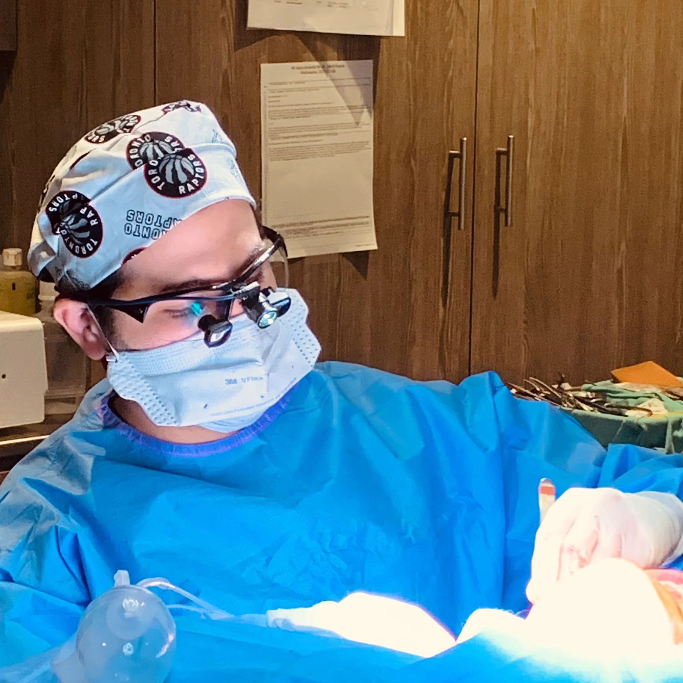A surgeon wearing a Toronto Raptors scrub cap, glasses, a surgical mask, and blue scrubs is performing surgery in an operating room with wooden cabinets in the background.