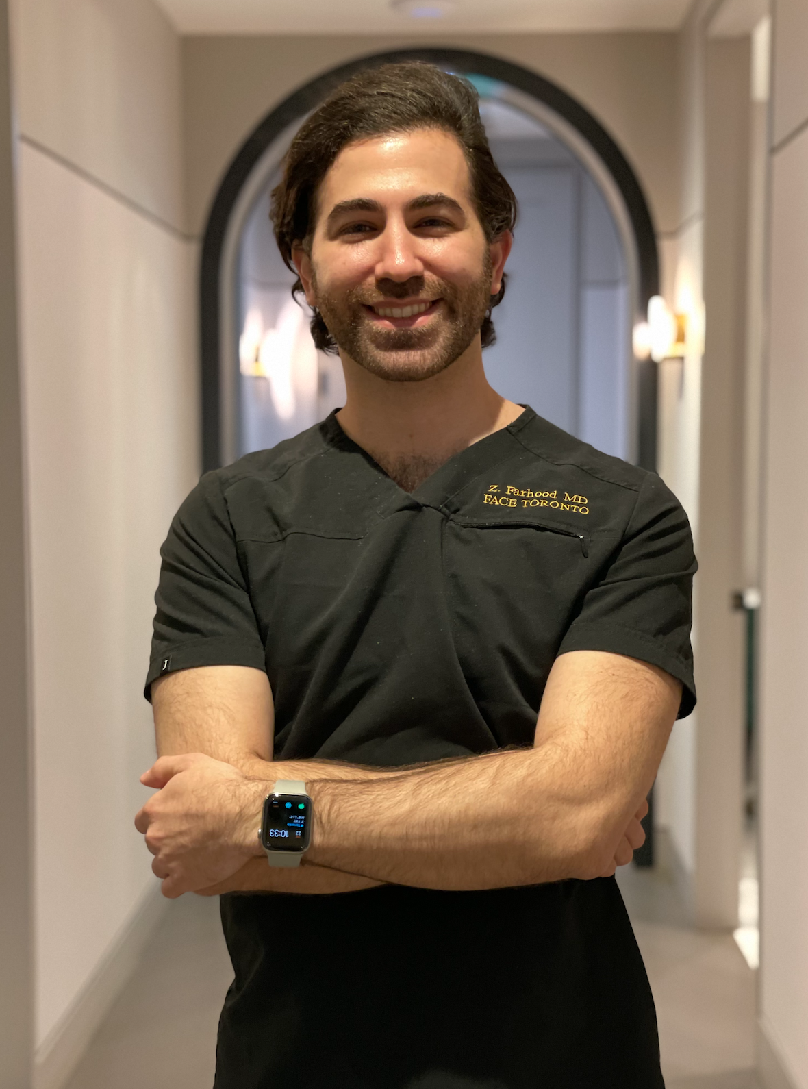 A smiling man with dark hair and a beard wearing black medical scrubs with gold lettering, standing in a corridor with his arms crossed and wearing a smartwatch.