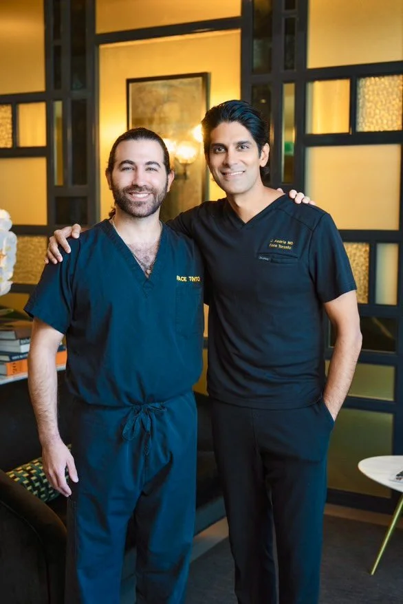 Two men in black medical scrubs smiling and posing together in an indoor setting with warm lighting and decorative wall panels.