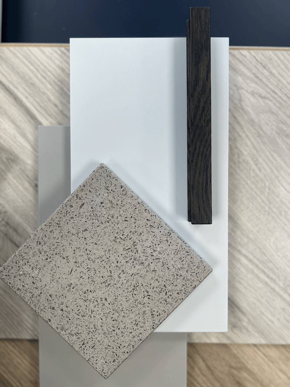 A sample board showcasing different material samples, including a speckled beige tile, a glossy white tile, a dark wood strip, and a wood grain surface background.
