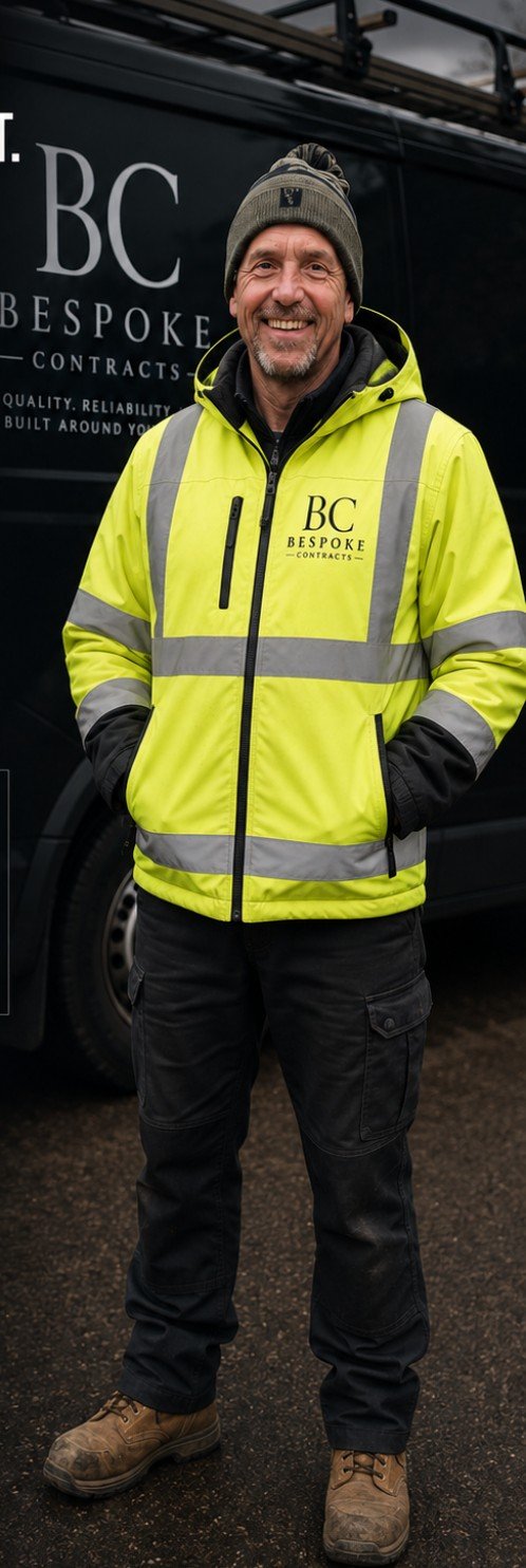 A smiling man wearing a gray beanie and a bright yellow safety jacket standing in front of a black work van with 'BC BESPOKE CONTRACTS' logo.