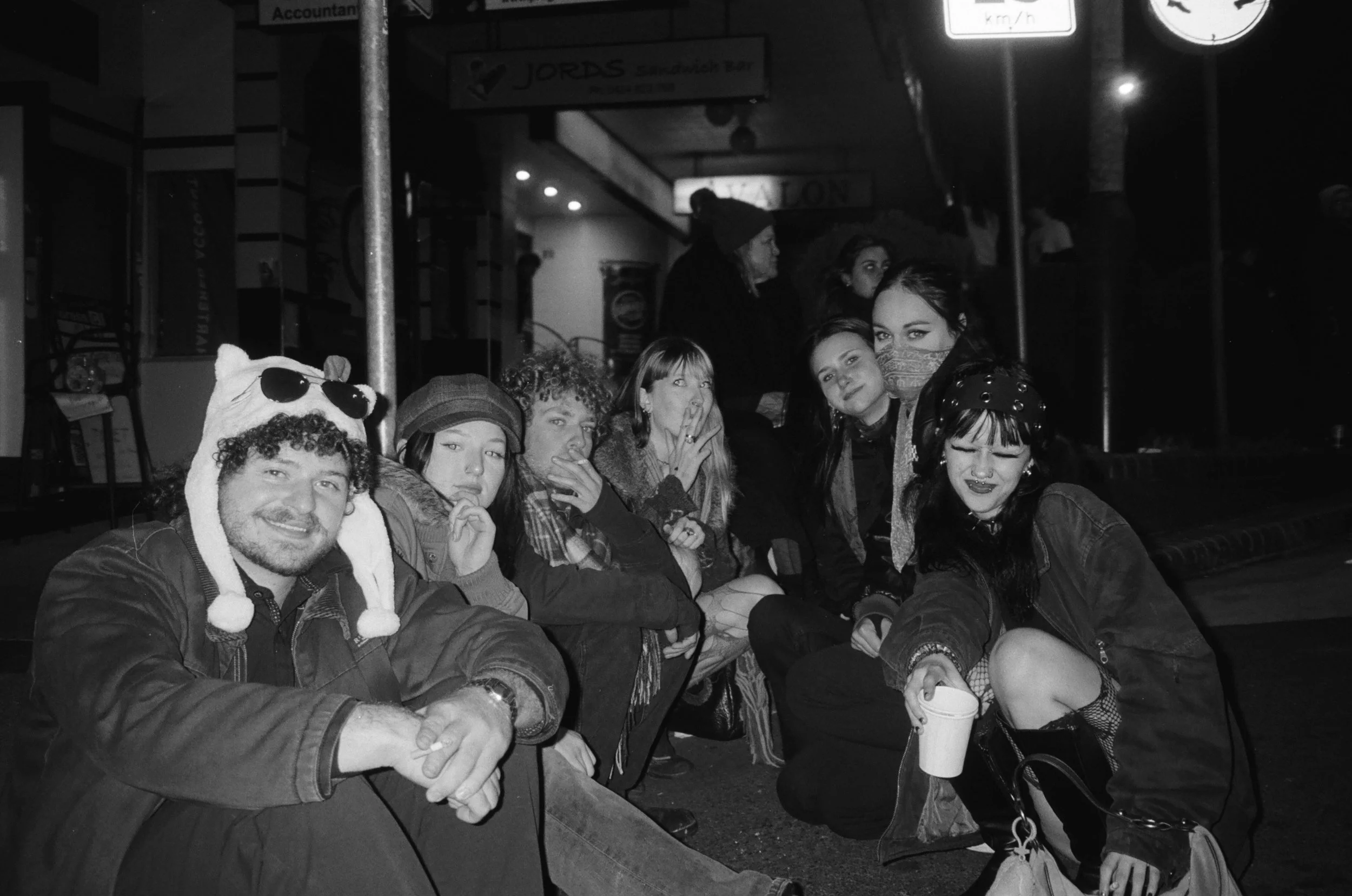A group of young people sitting close together on a bench outside at night. They are casually dressed, some with accessories like hats and face masks. Some are making peace signs or smiling at the camera. The background shows illuminated signs and st