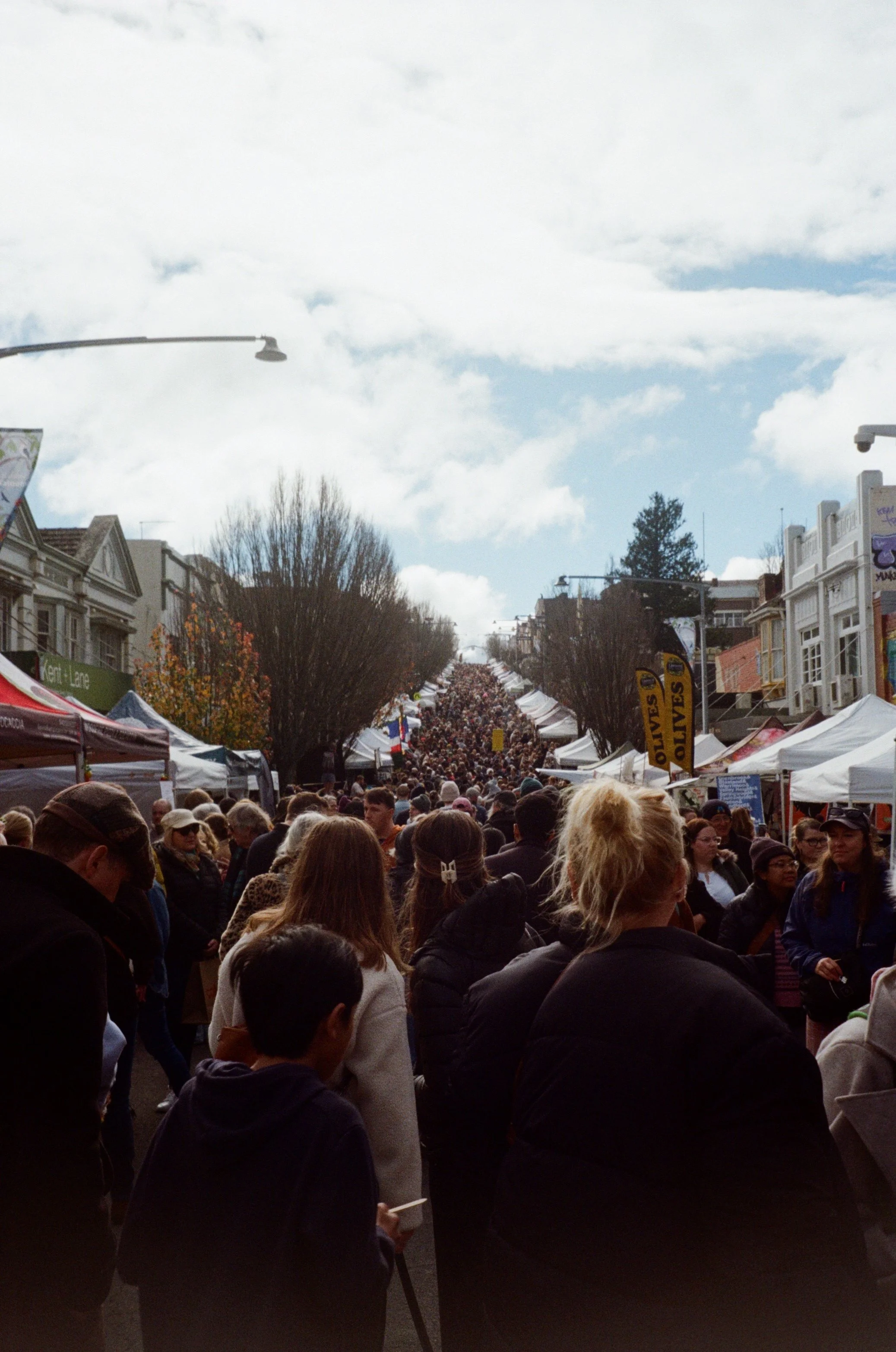 Crowd of people walking on a busy street during a festival or market, with tents on both sides, under a partly cloudy sky, and city buildings in the background.