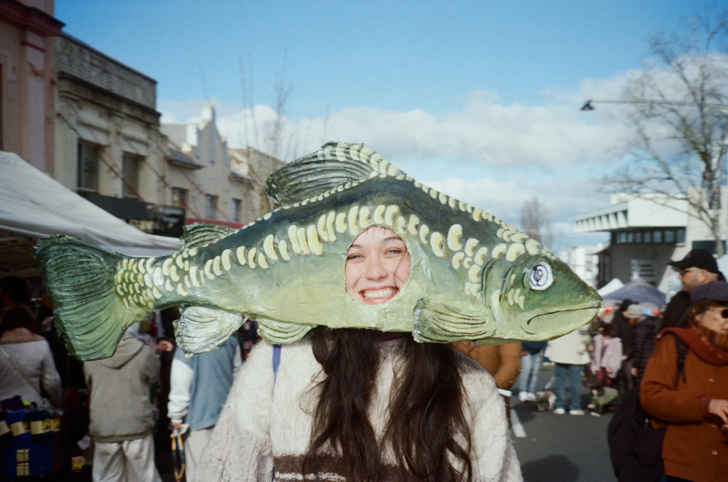 A woman wearing a large green paper mache fish headpiece at a winter festival among many people and on a crowded street