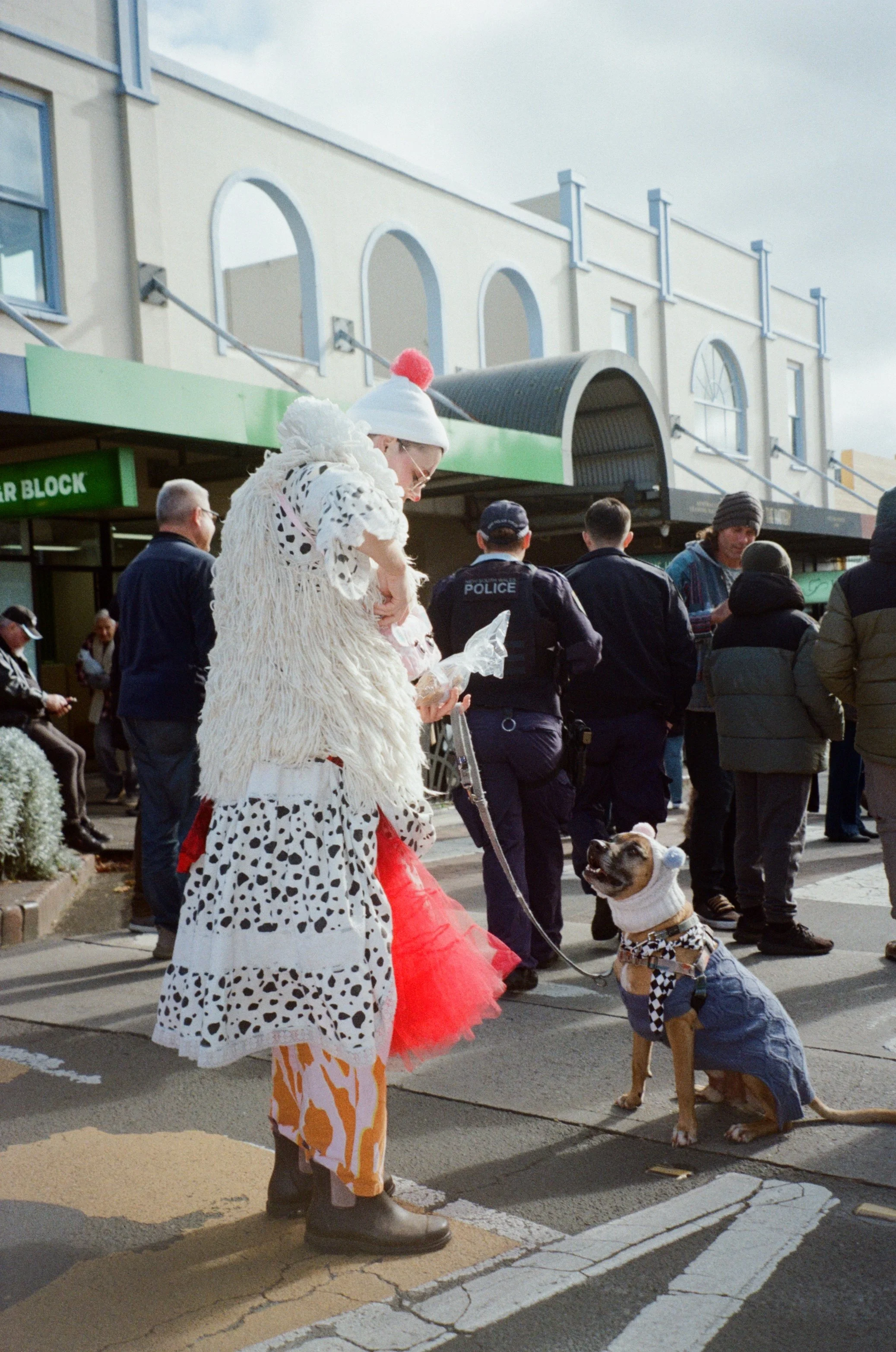 A person dressed in a sheep costume with a white hat and red pompom adjusts their clothing while another dressed as a dog in a plaid vest with a bow tie looks up at them. Several people are gathered on the street in the background, including police o
