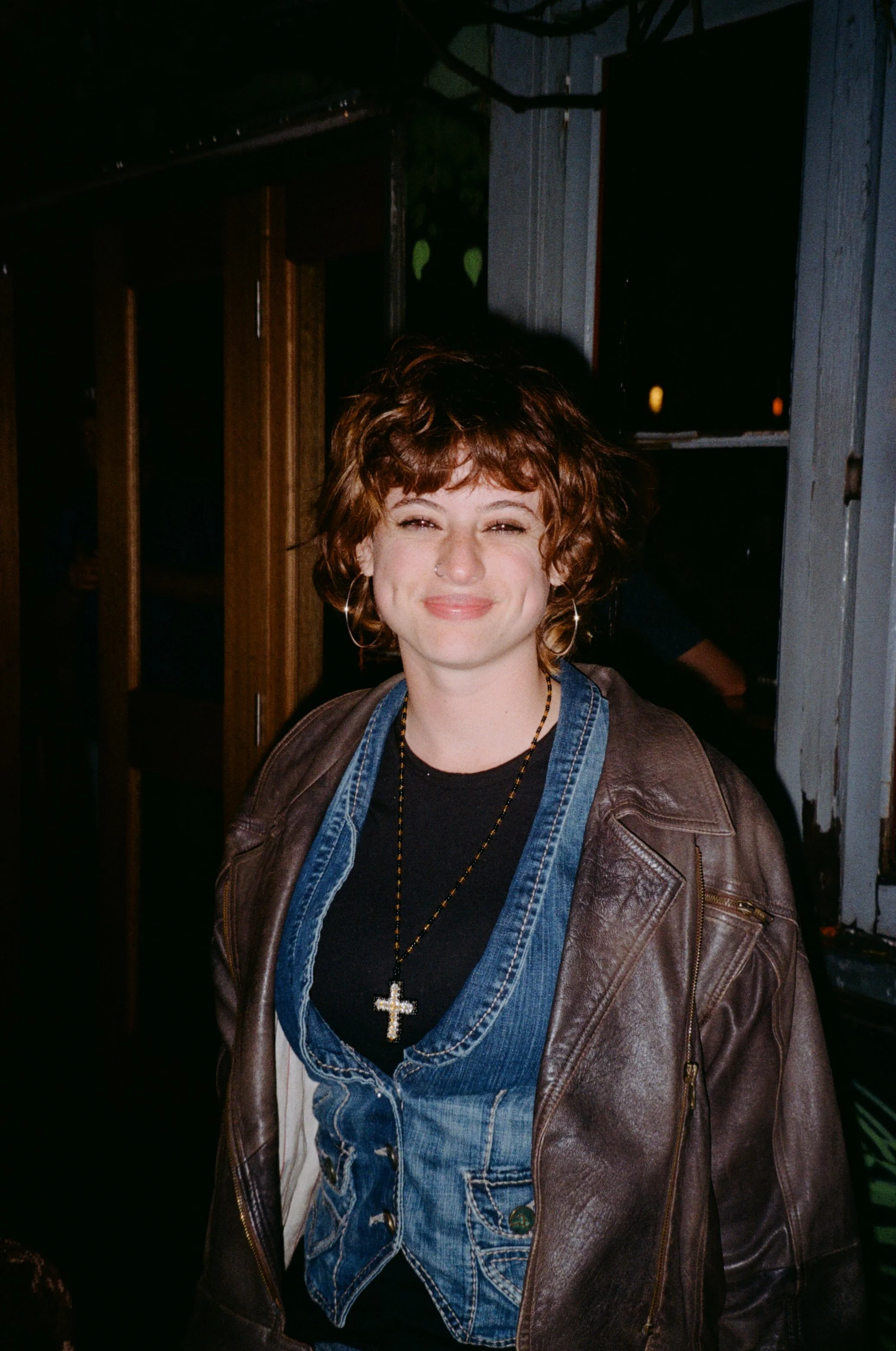 A woman with curly brown hair smiling at night, wearing a black shirt, denim vest, and brown leather jacket, with a cross necklace in an outdoor bar setting.