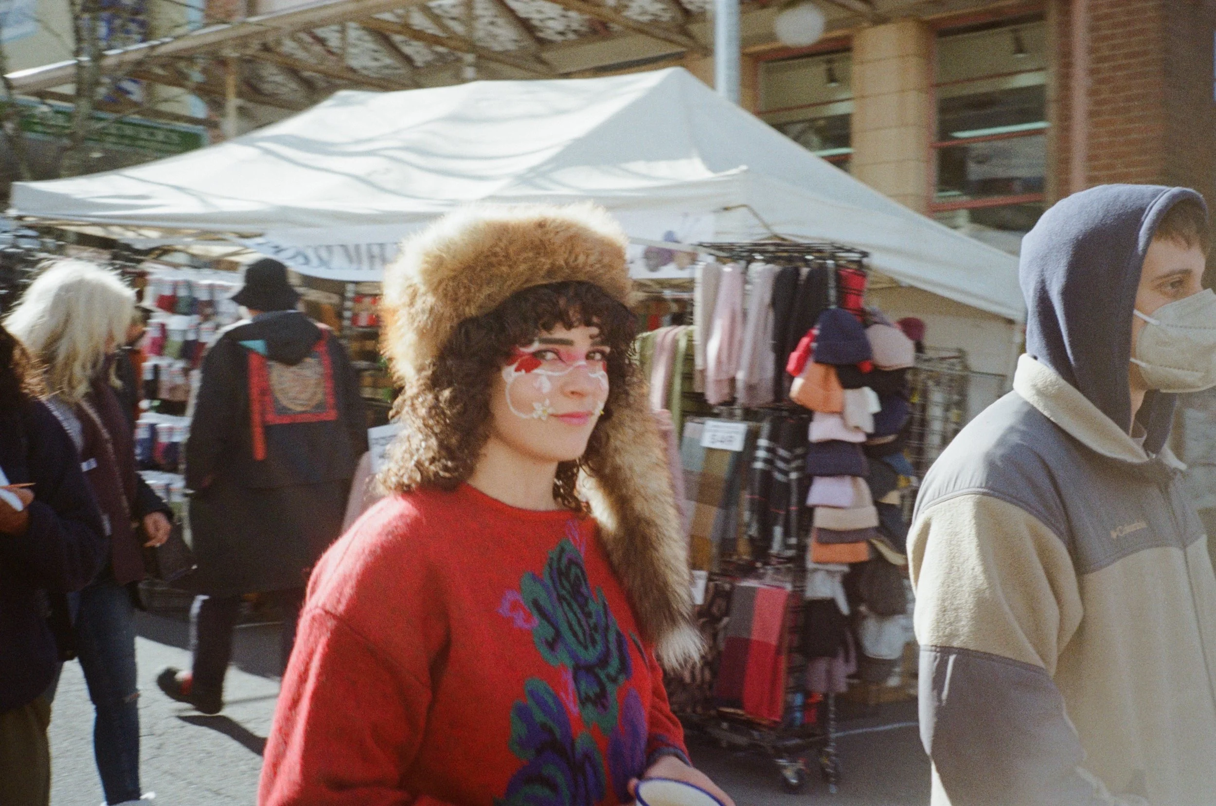A woman with curly hair, painted face, and wearing a fur hat and red sweater is walking outdoors at a market, with people and market stalls in the background.