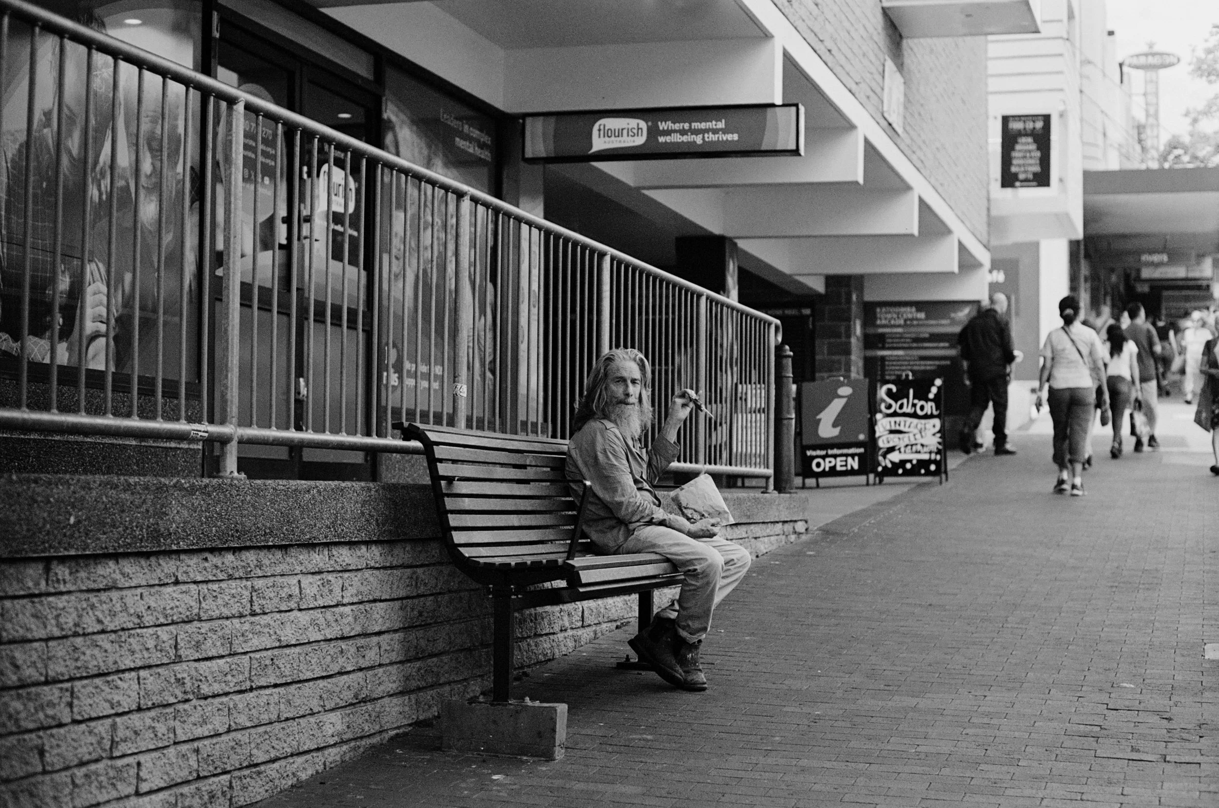 A man sitting on a bench outside a building, holding papers or a bag, with pedestrians walking by on the street. There are signs and storefronts visible, including a sign that says 'flourish' and another advertising 'Salon VINTAGE.' The scene is in b