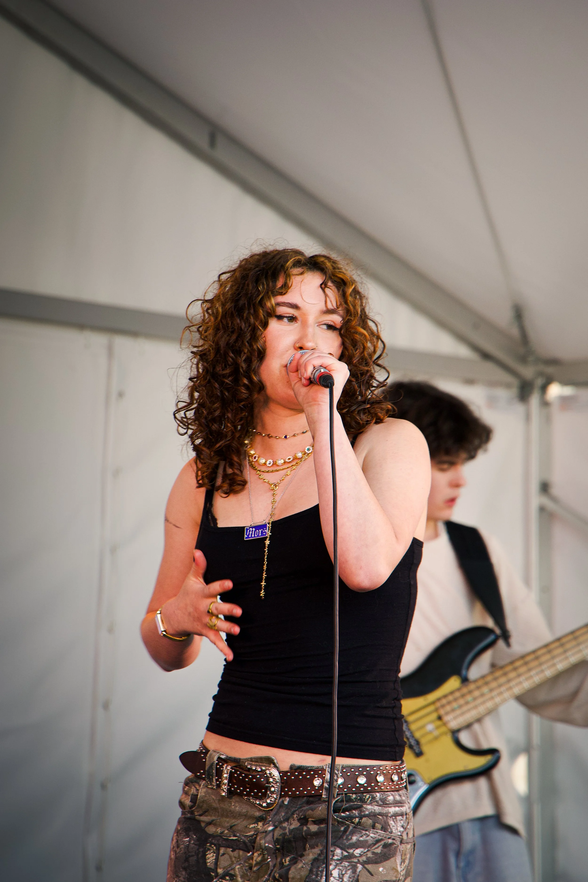 A young woman with curly hair singing into a microphone, wearing a black tank top, layered necklaces, and camouflage pants topped with a studded belt. In the background, a person with long hair playing an electric guitar.