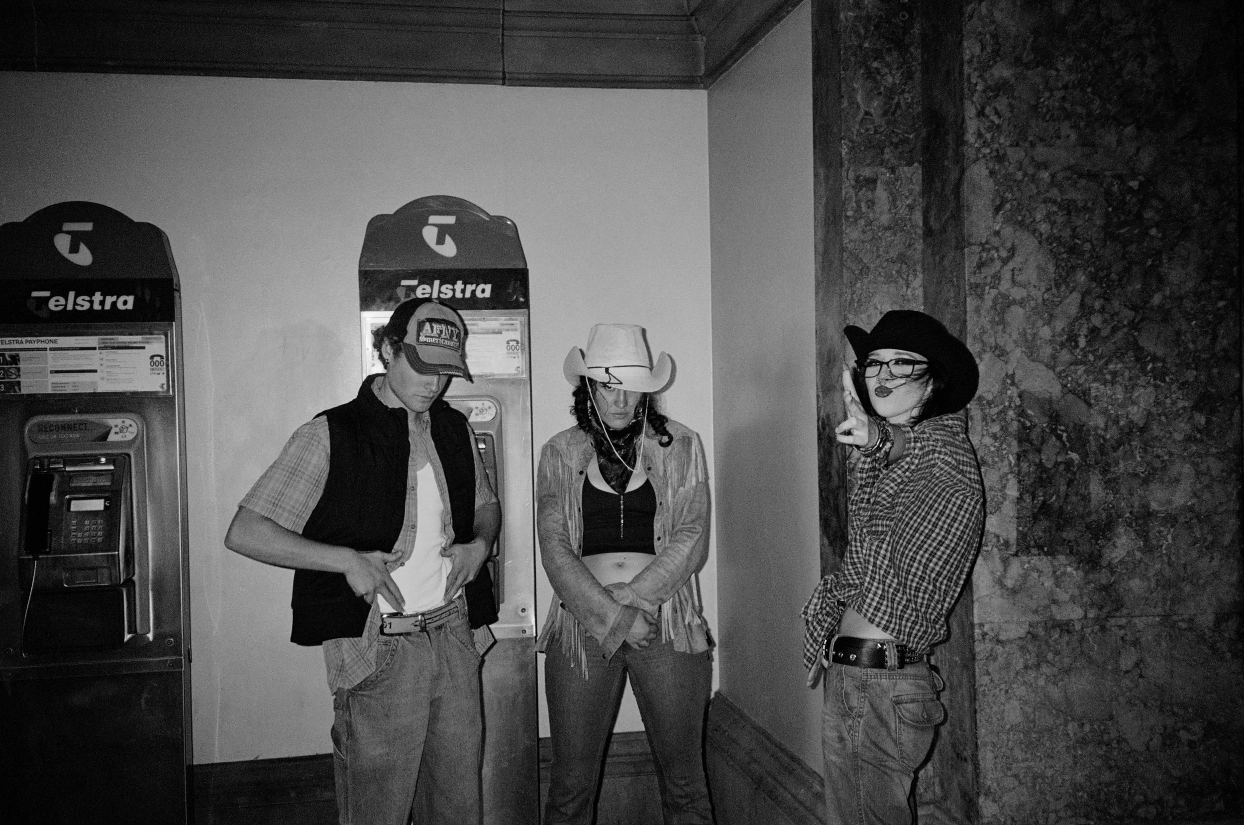 Four people standing in front of two payphones, with a stone wall on the right side. They are dressed in casual and Western-style outfits, with one person wearing a cowboy hat and another wearing a wide-brimmed hat. The image is in black and white.