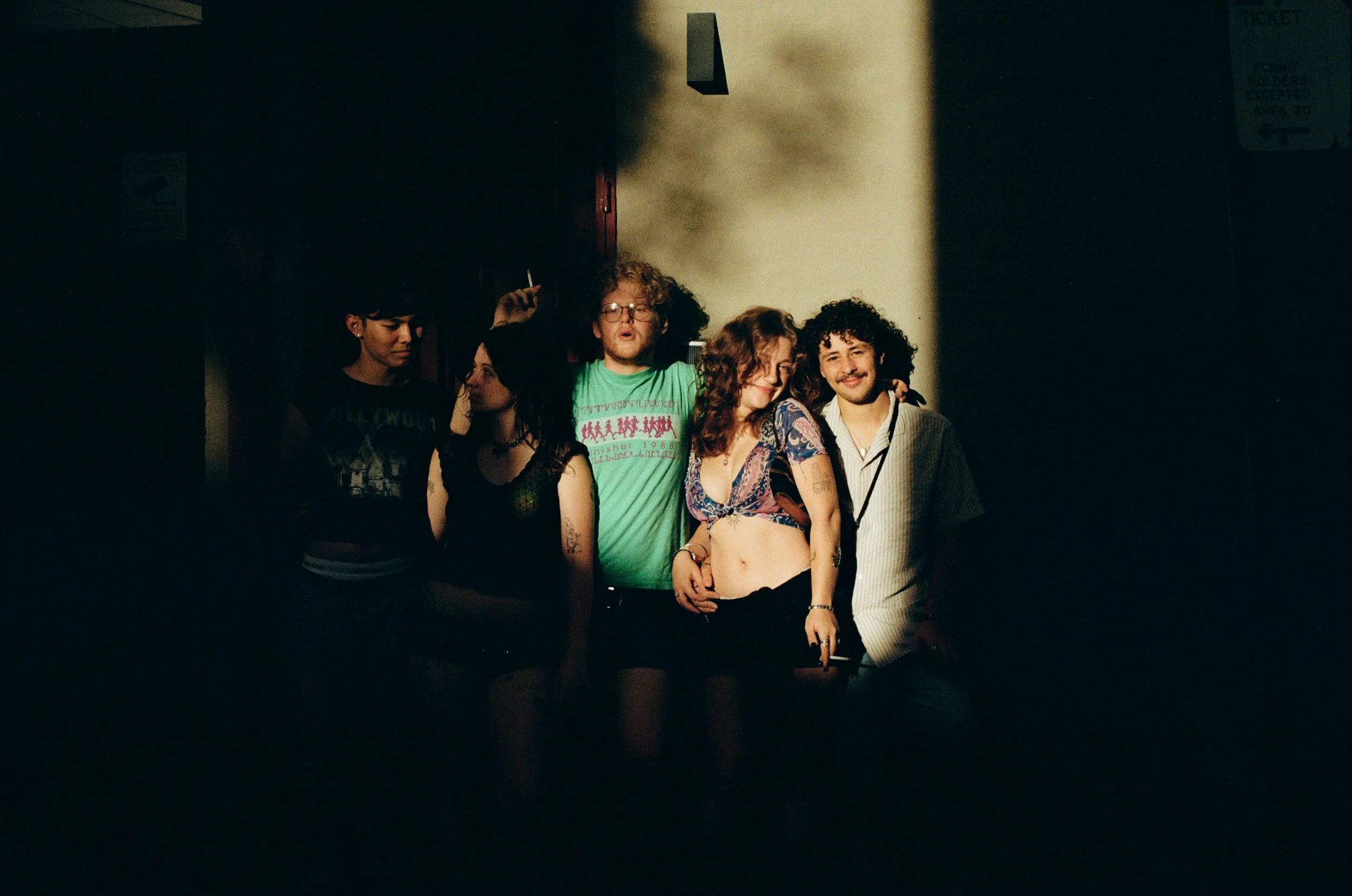 A group of five friends, three women and two men, standing together on a dimly lit street pathway. They are celebrating and smiling, with one man holding a cigarette in his hand. The group appears relaxed and happy.